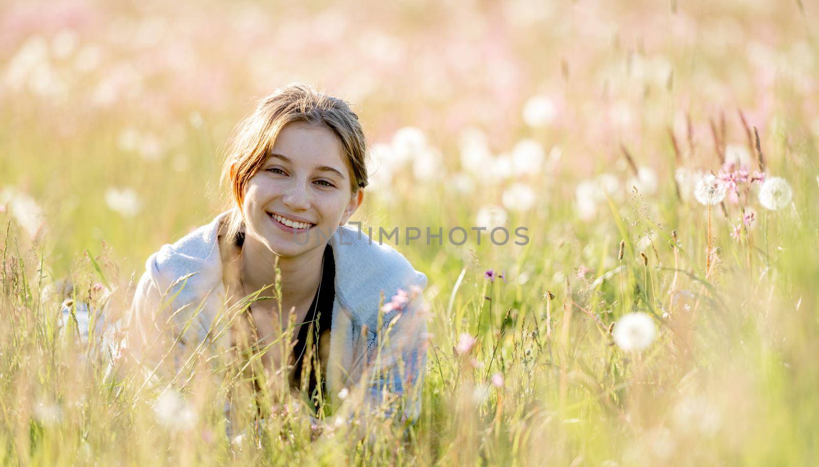 Pretty girl teenager sitting in field with dandelions and smiling outdoors. Beautiful young female person at nature with flowers