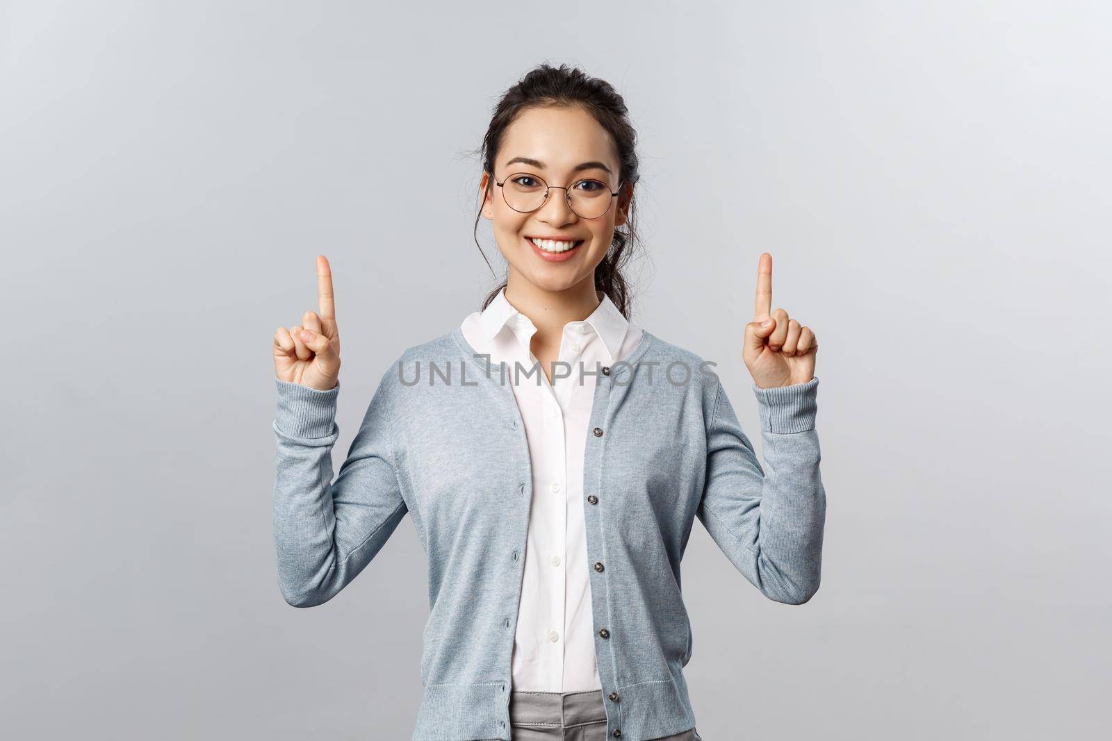 Friendly cheerful asian girl in glasses, showing product or company banner, pointing fingers up inviting check-up event news, info about advertisement, smiling adorable at camera, grey background.