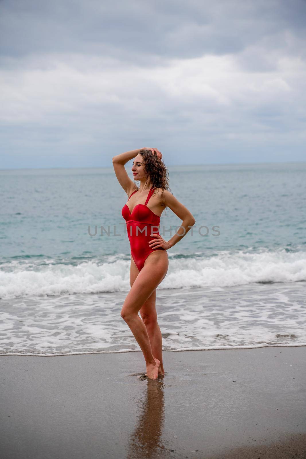 A beautiful and sexy brunette in a red swimsuit on a pebble beach, Running along the shore in the foam of the waves by Matiunina