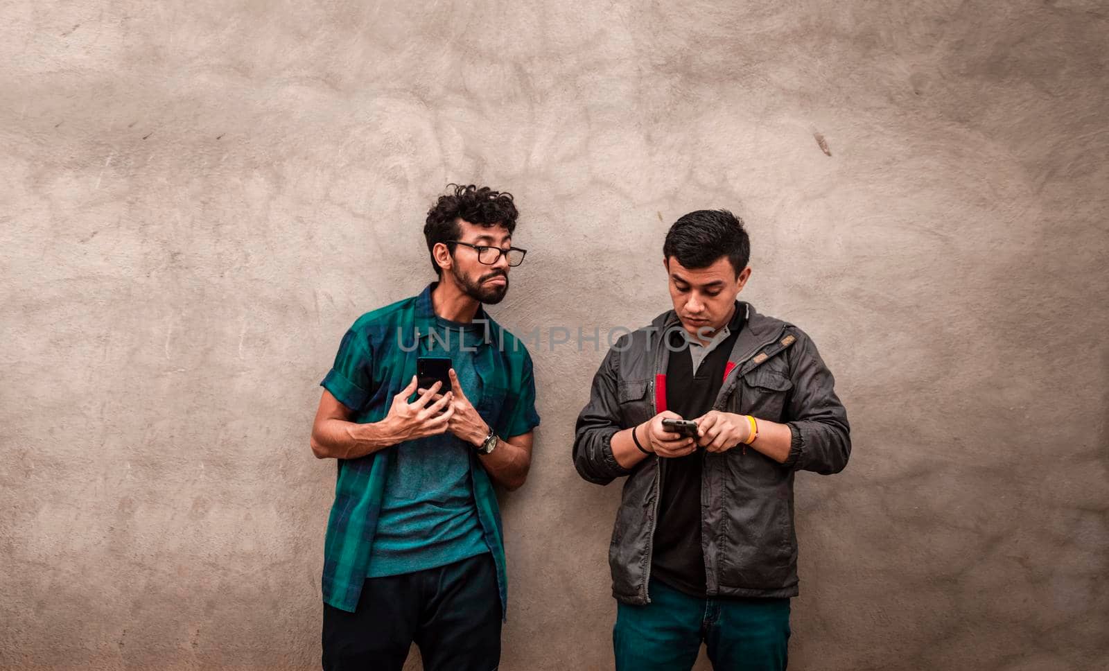 Young latin man spying on another cell phone, man showing his cell phone to another guy