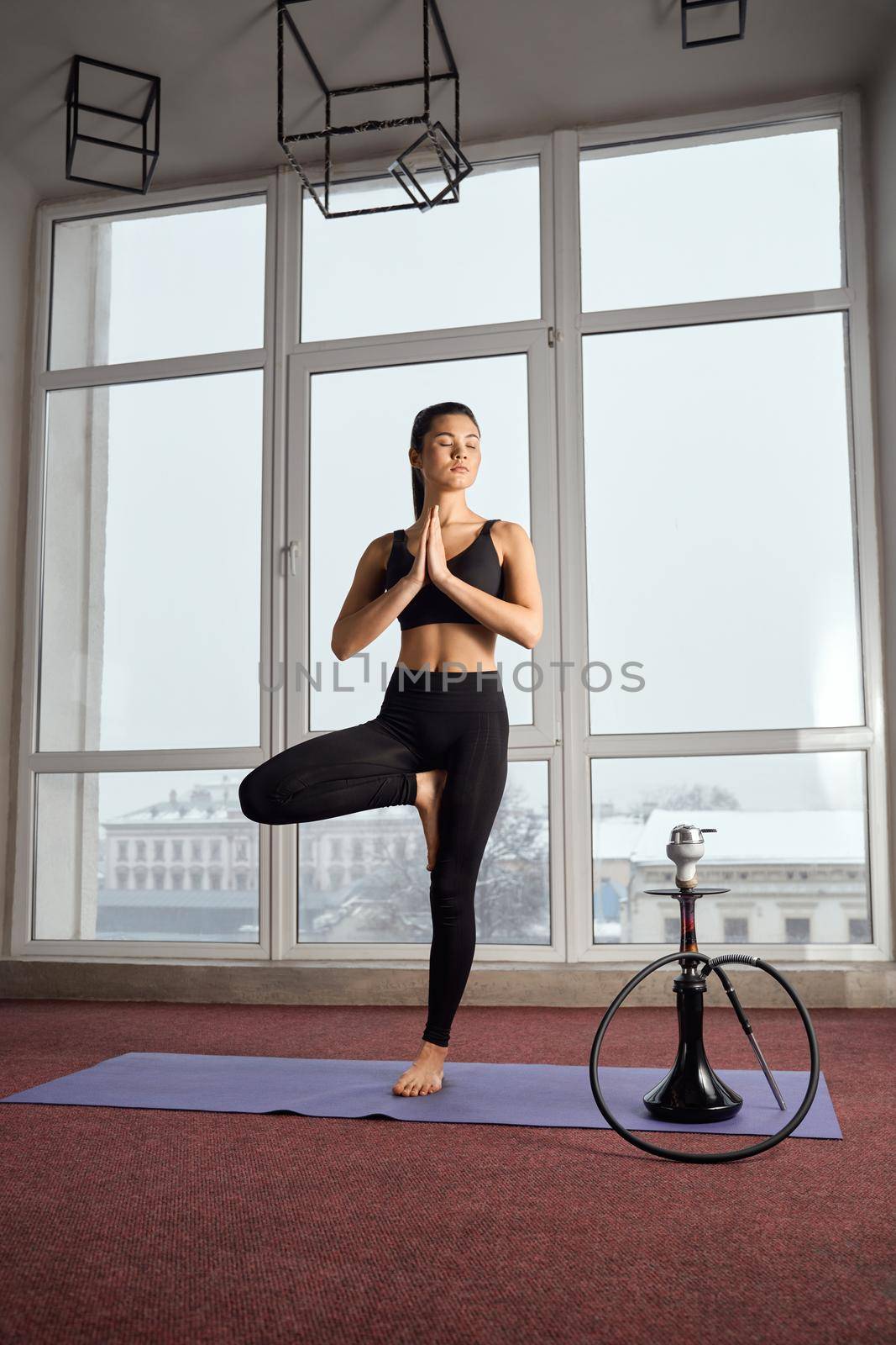 Side view of sporty girl in black sportswear standing in tree pose of yoga with closed eyes on mat. Barefoot lady practicing yoga in specious room, with hookah on background. Concept of yoga practice.