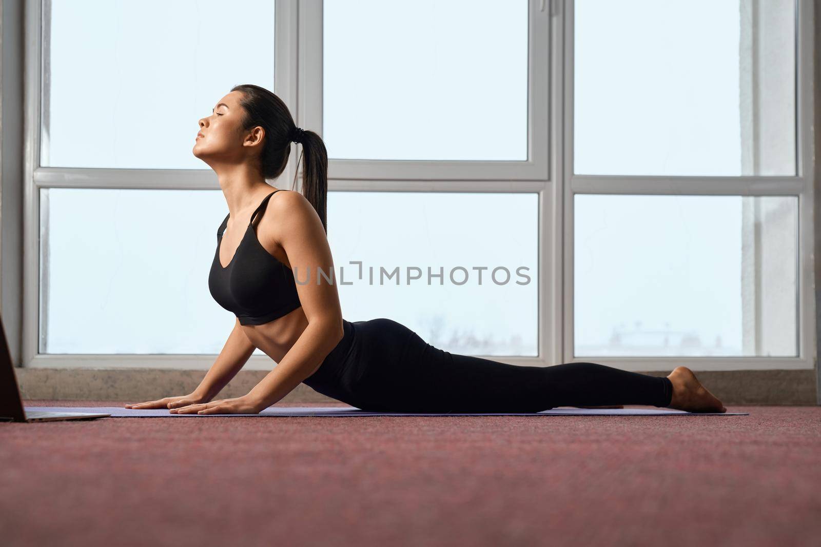 Side view of attractive woman stretched in upward dog pose of yoga while having yoga class. Fit lady with closed eyes laying in yoga asana on blue mat, relaxing in calm atmosphere. Concept of yoga.