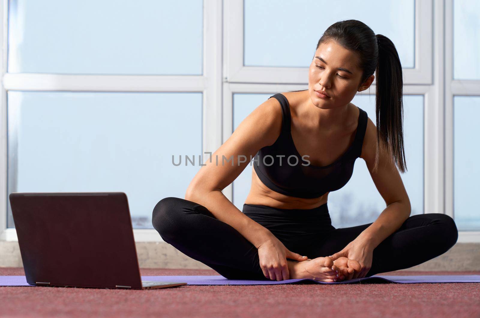 Front view of young barefoot woman having online yoga class at home. Beautiful girl exercising in butterfly II yoga pose, looking at laptop during interactive classes. Concept of yoga practicing.