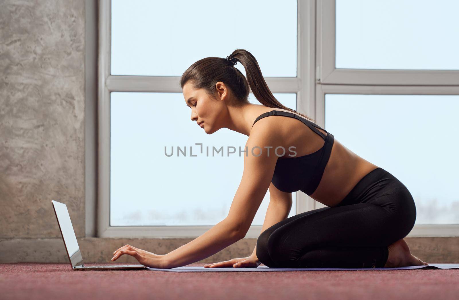 Side view of fit pretty woman doing yoga, looking at laptop at home. Attractive lady in sportswear sitting on knees, during online classes at home with computer in front of women. Concept of yoga.