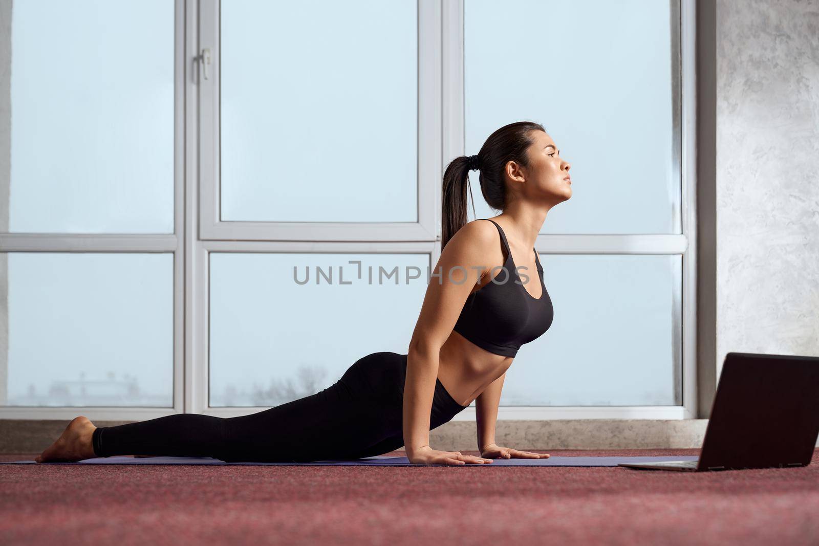 Side view of slim female in sportswear standing in upward-facing dog pose while having online yoga class. Young caucasian barefoot girl practicing yoga on mat along in spacious room. Concept of yoga.