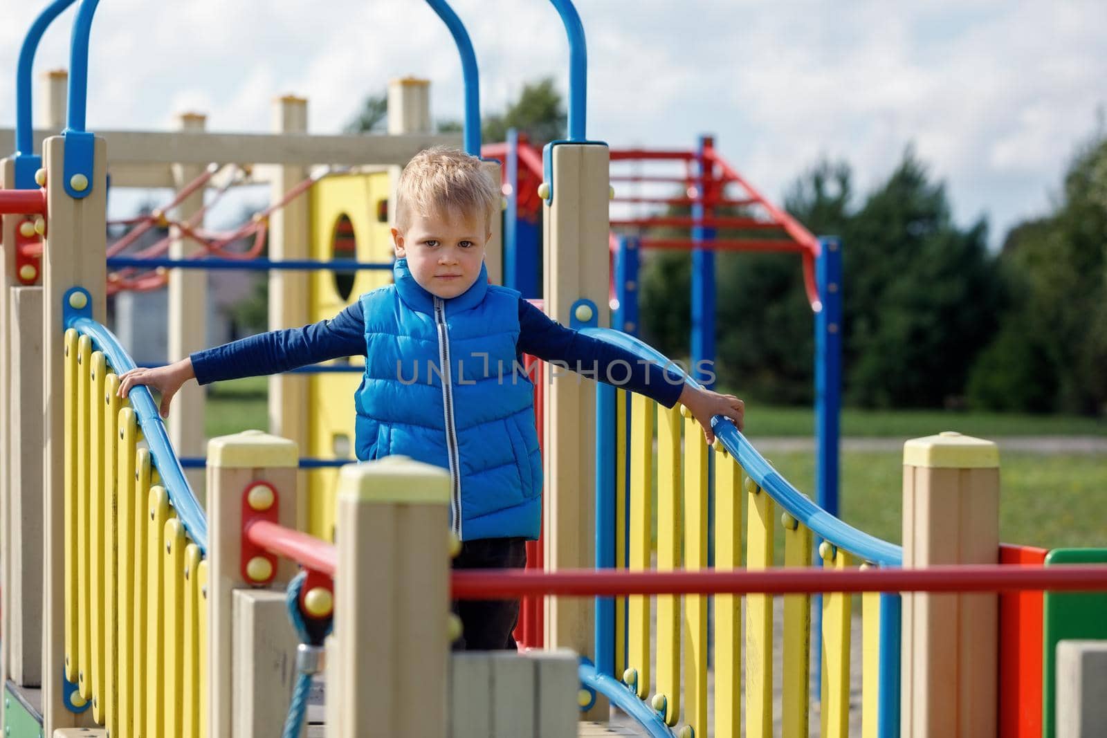 A little boy wearing a blue vest walks on a wavy bridge on a colorful outdoor playground. by Lincikas