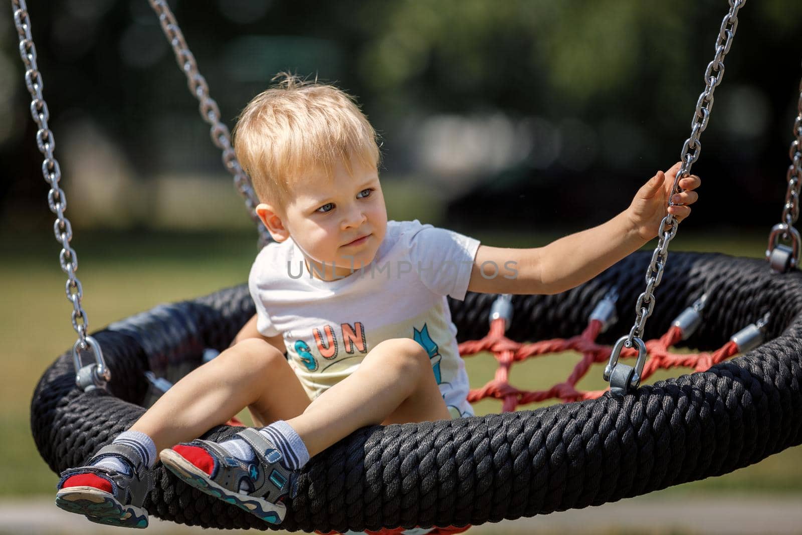 A cheerful little boy swings in the park on a swinging rope swing. Round black and red seat for children's swing.