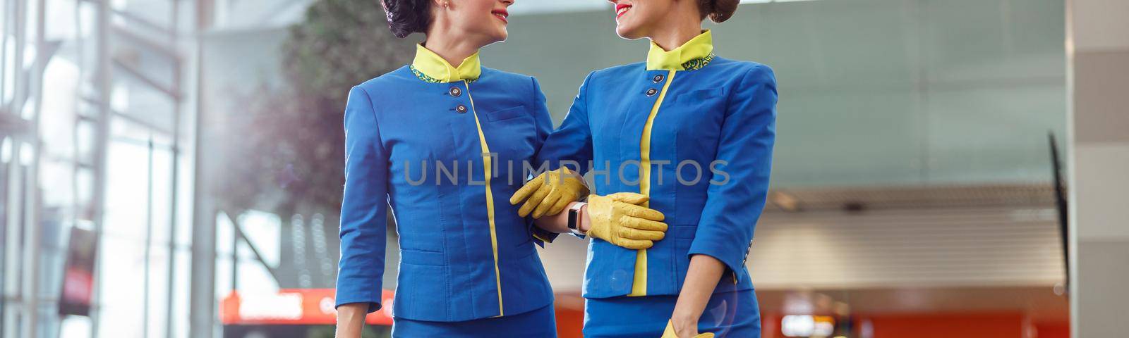 Two cheerful women stewardesses in aviation air hostess uniform looking at each other and smiling while holding travel bags
