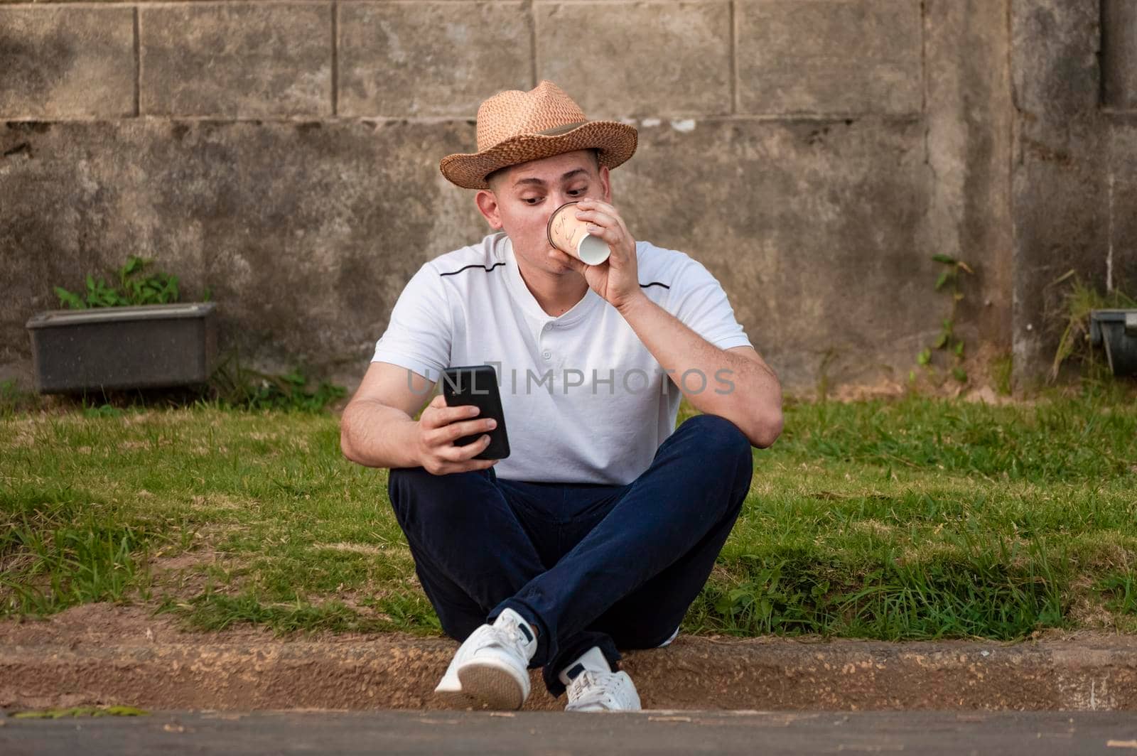 Young man sitting on sidewalk checking phone, man drinking coffee while checking his phone