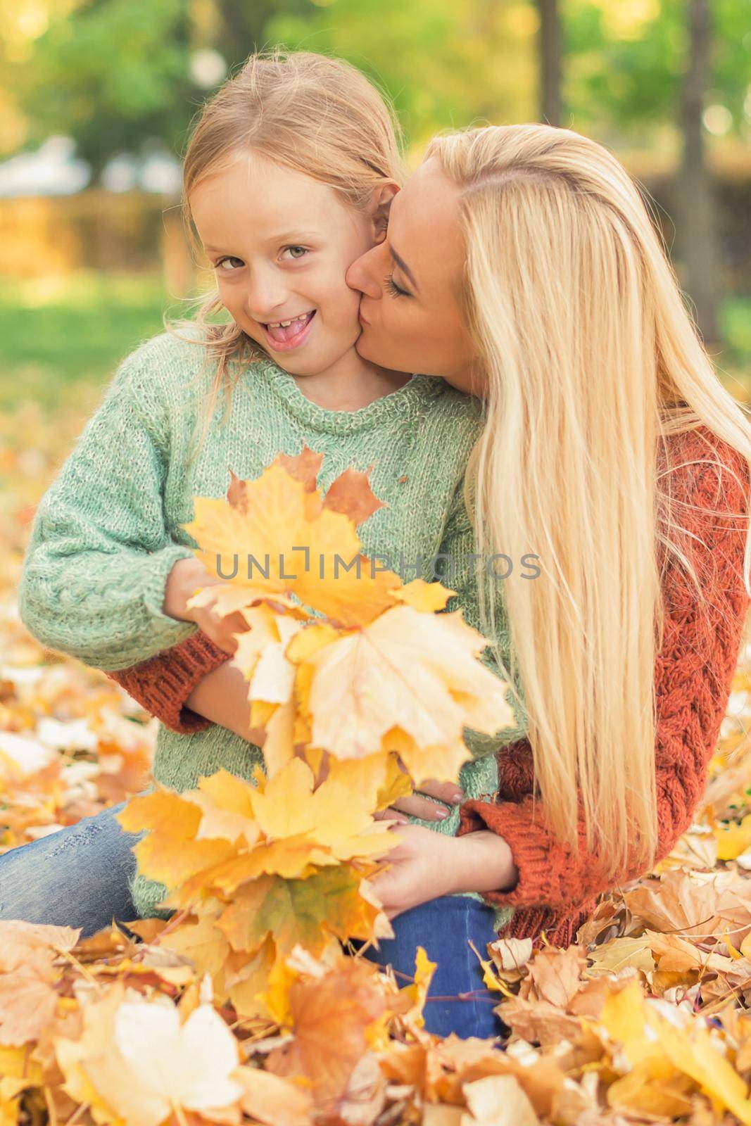 Happy young caucasian mother and little daughter holding autumn yellow leaves sitting and kissing at the park