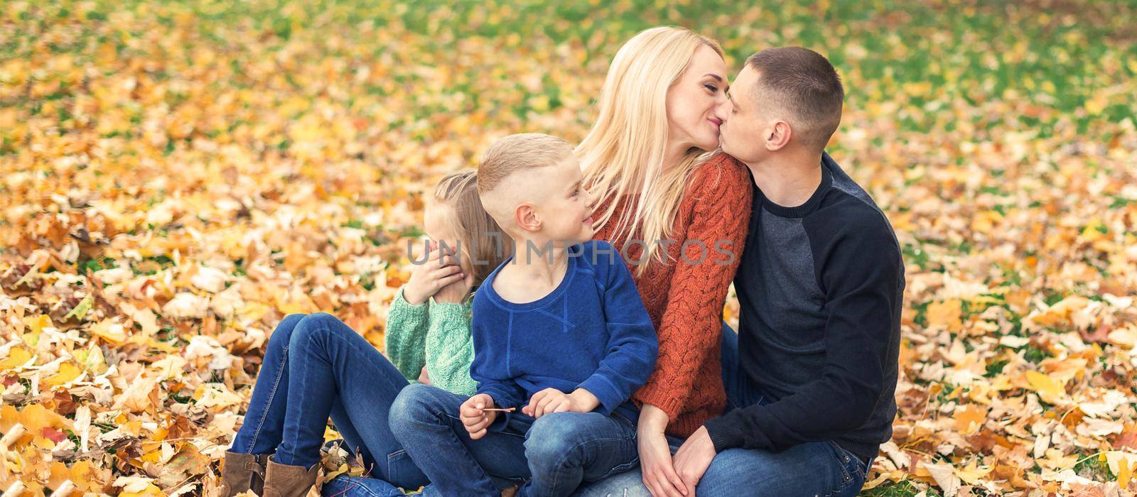 Portrait of young family sitting in autumn leaves. Parents kissing and sitting with children in the autumn park