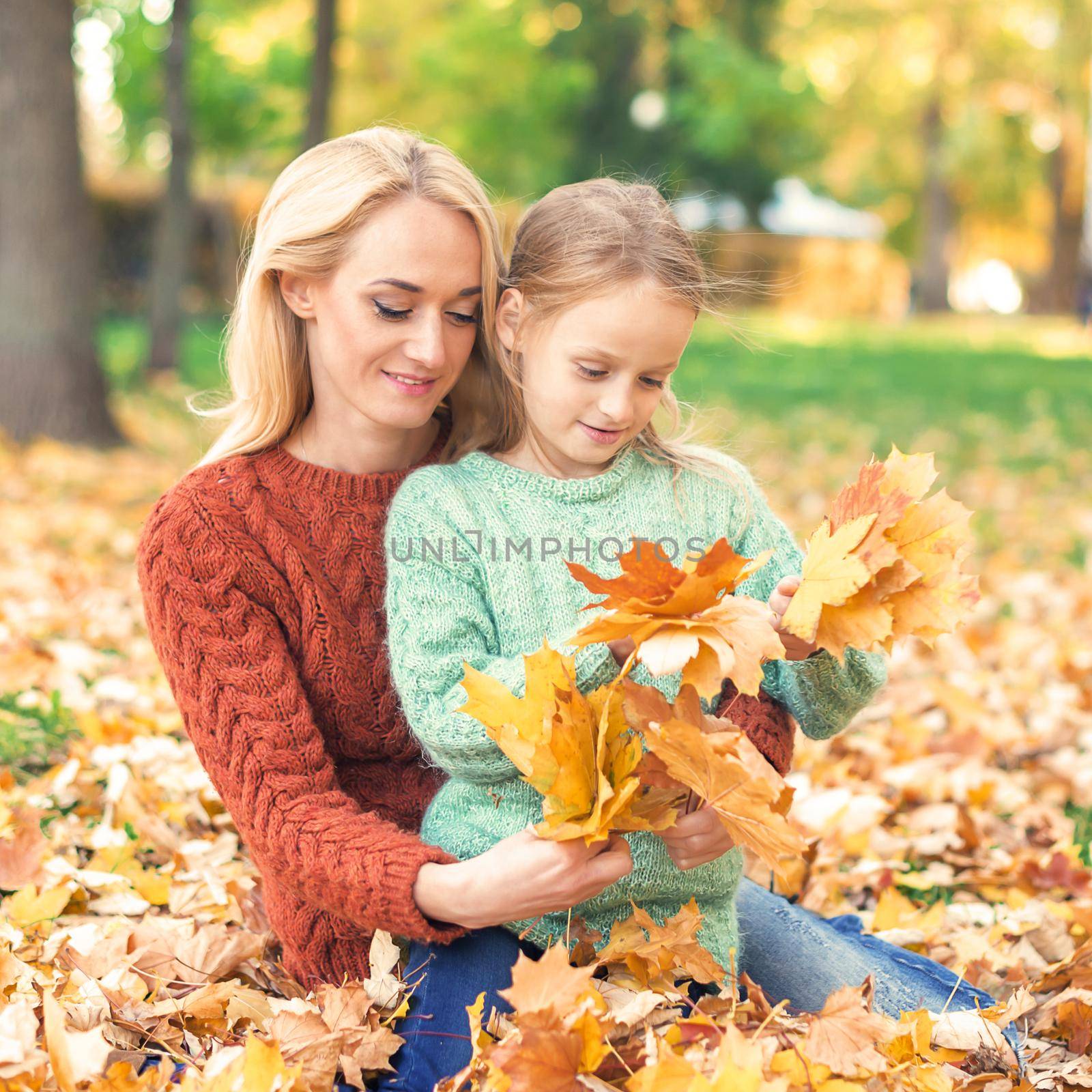 Happy young caucasian woman and little girl holding autumn yellow leaves sitting at the park