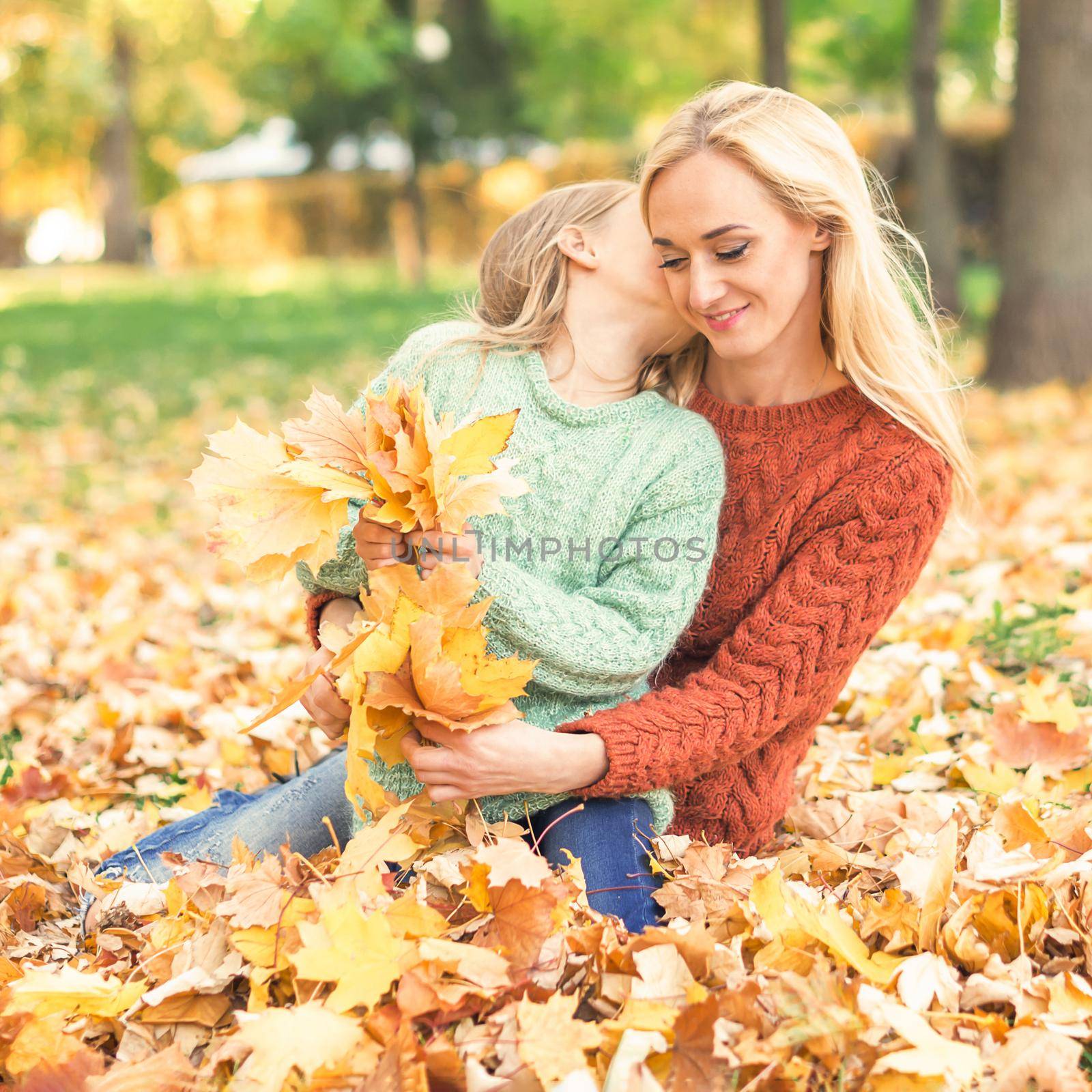 Happy young caucasian mother and little daughter holding autumn yellow leaves sitting and kissing at the park