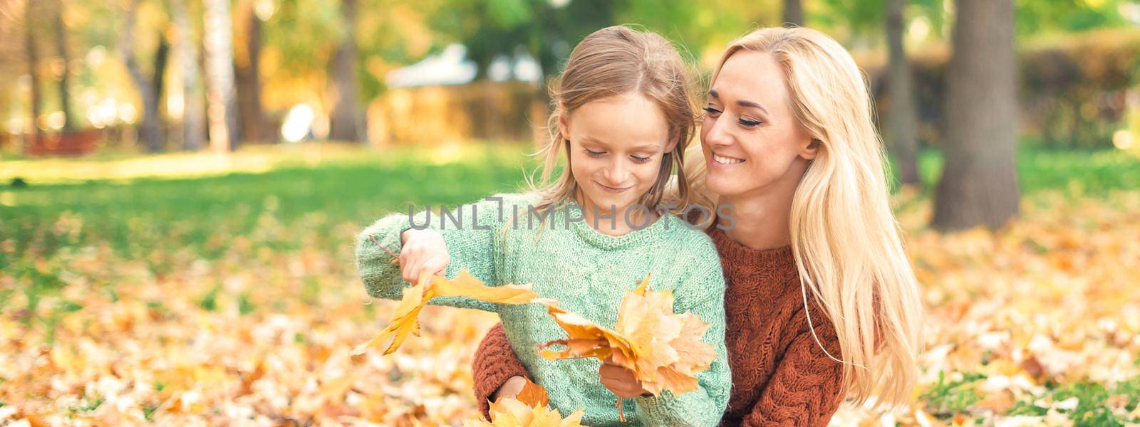 Happy young caucasian mother and little daughter holding autumn yellow leaves sitting at the park