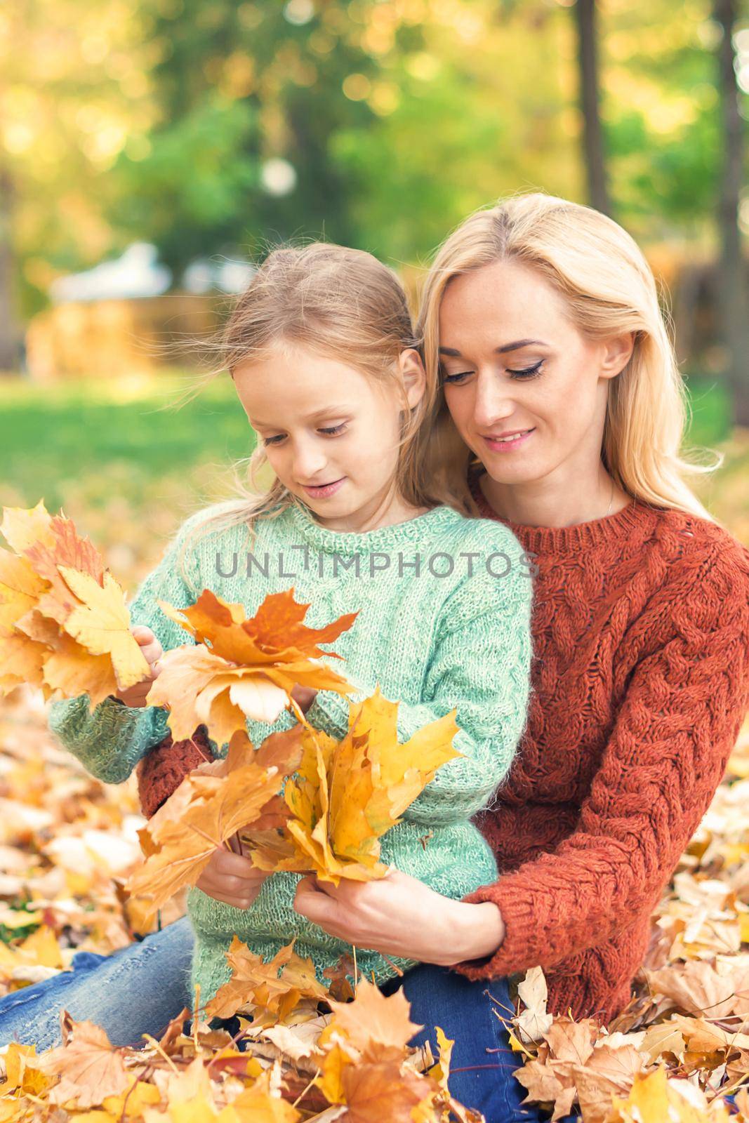 Happy young caucasian woman and little girl holding autumn yellow leaves sitting at the park