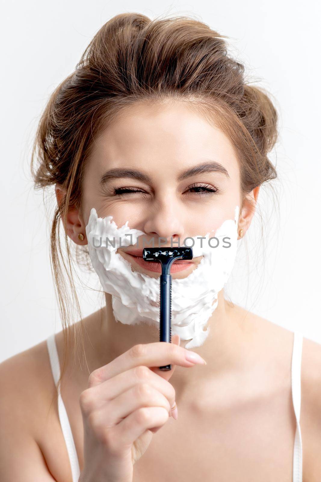 Beautiful young caucasian woman shaving her face by razor on white background. Pretty woman with shaving foam on her face