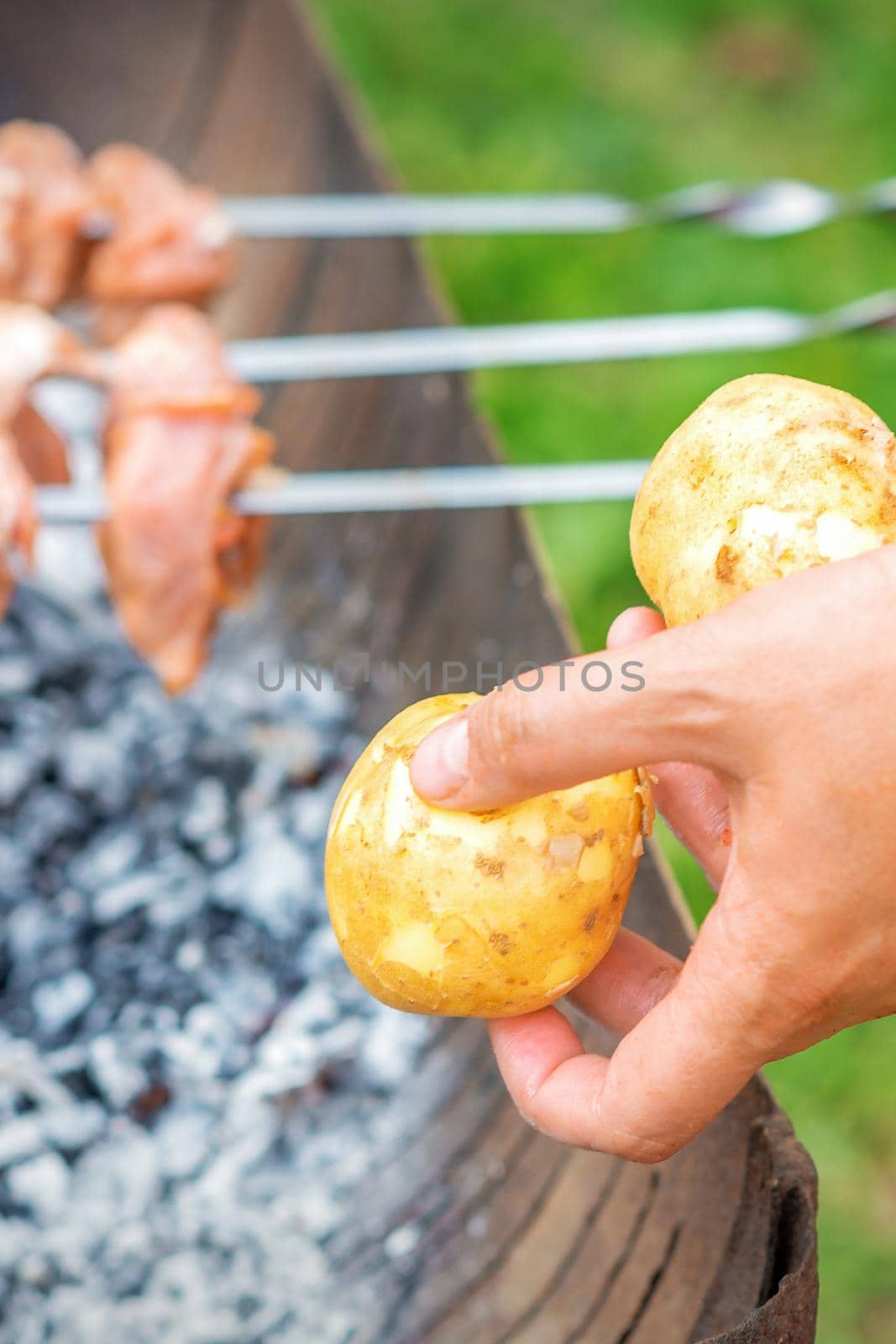 Hands of man prepares barbecue meat with potatoes on skewer by grill on fire outdoors. Concept of lifestyle rustic food preparation