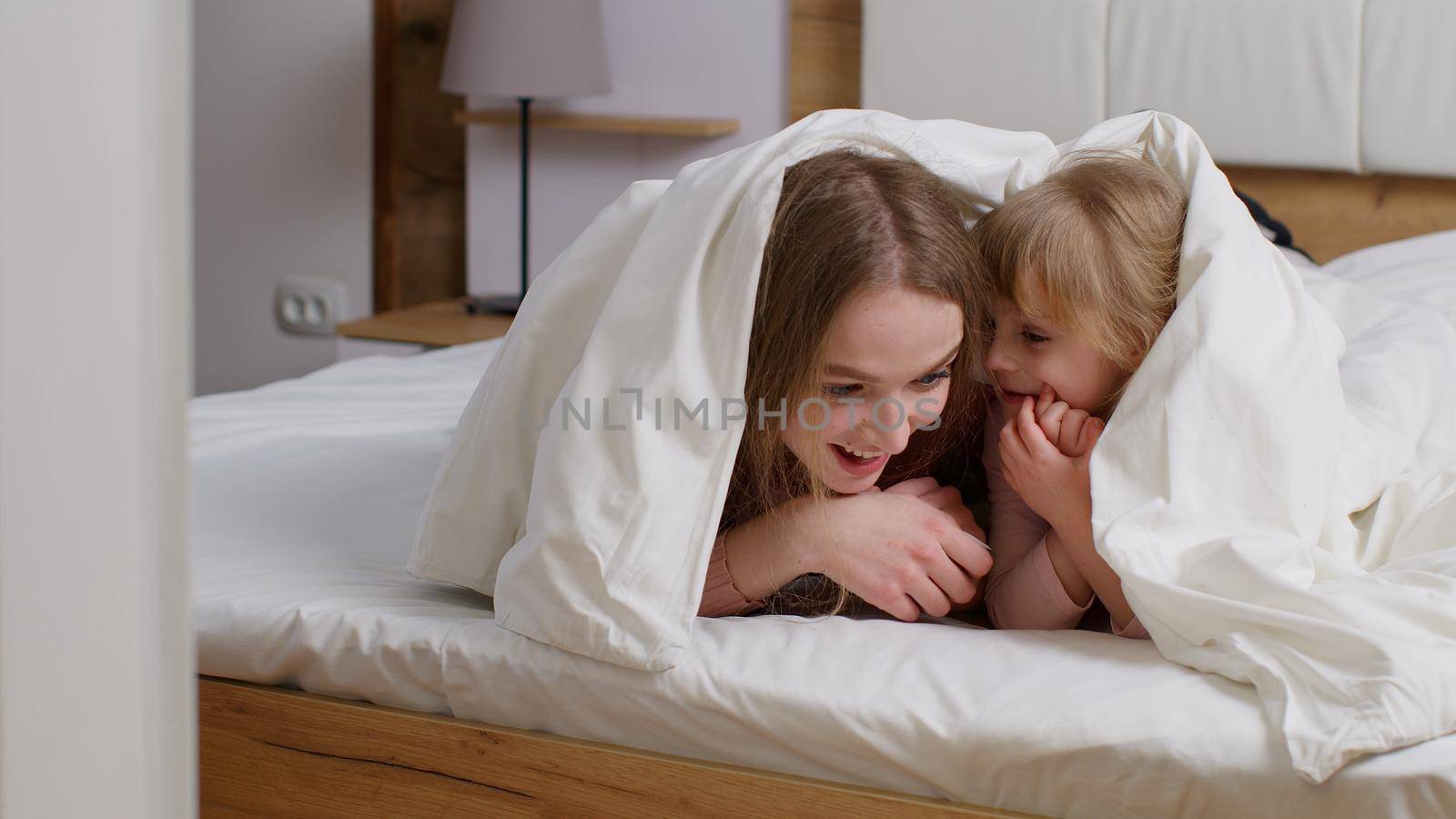 Happy family of mother with daughter girl lying on bed under duvet blanket, telling secrets to each other, having good time, smilling in bedroom. Parent and kid leisure hobbies activities together