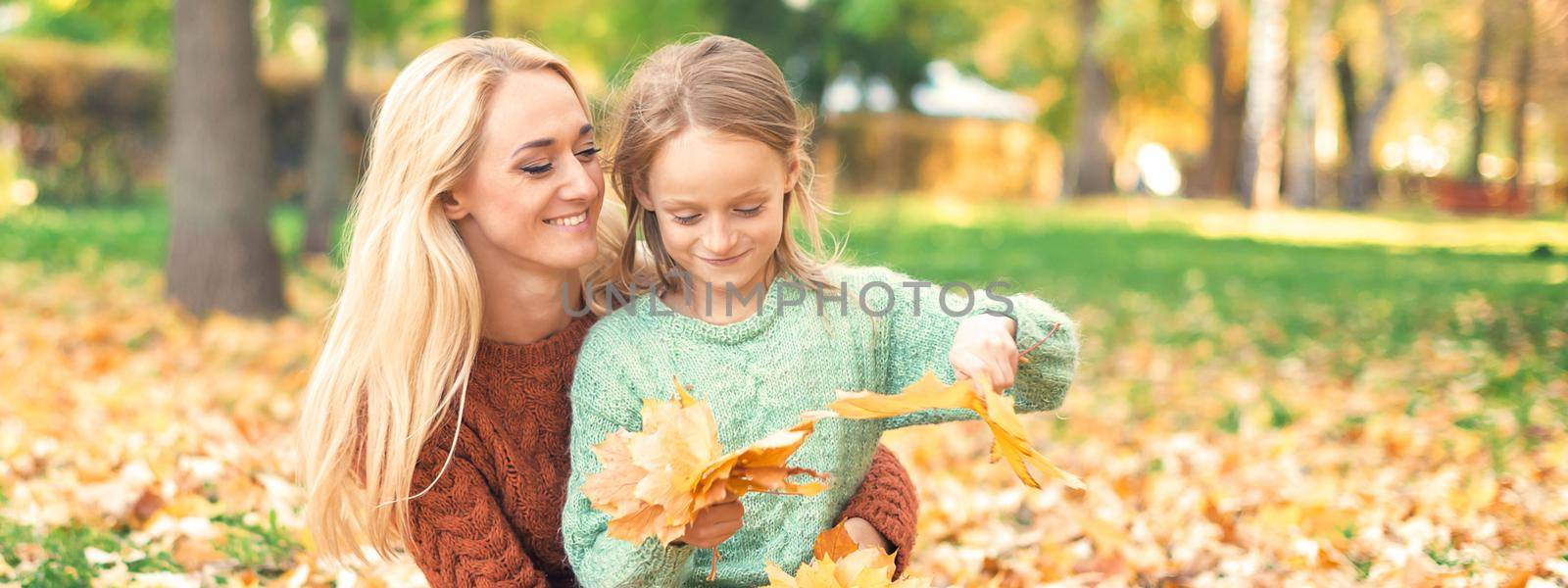 Happy young caucasian mother and little daughter holding autumn yellow leaves sitting at the park