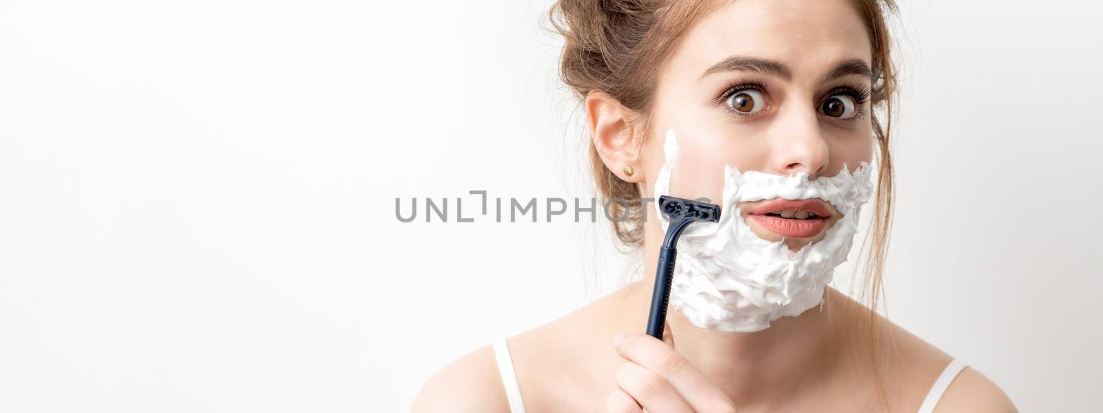 Beautiful young caucasian woman shaving her face by razor on white background. Pretty woman with shaving foam on her face