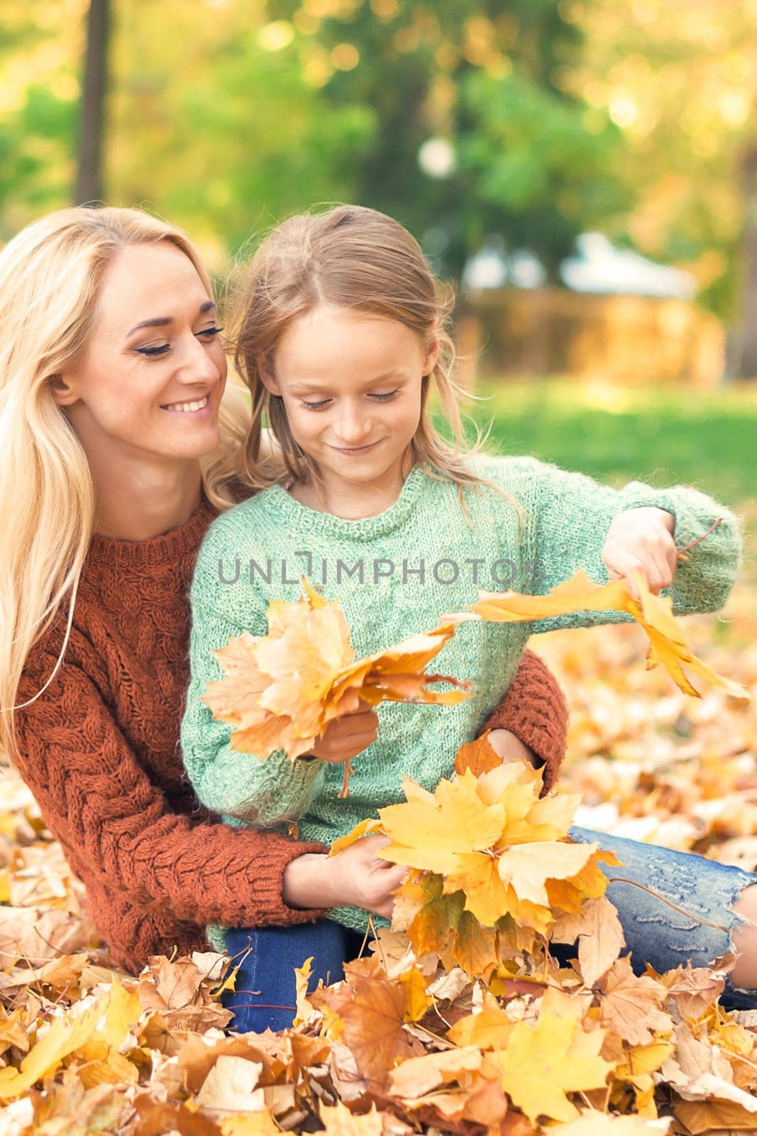 Happy young caucasian mother and little daughter holding autumn yellow leaves sitting at the park