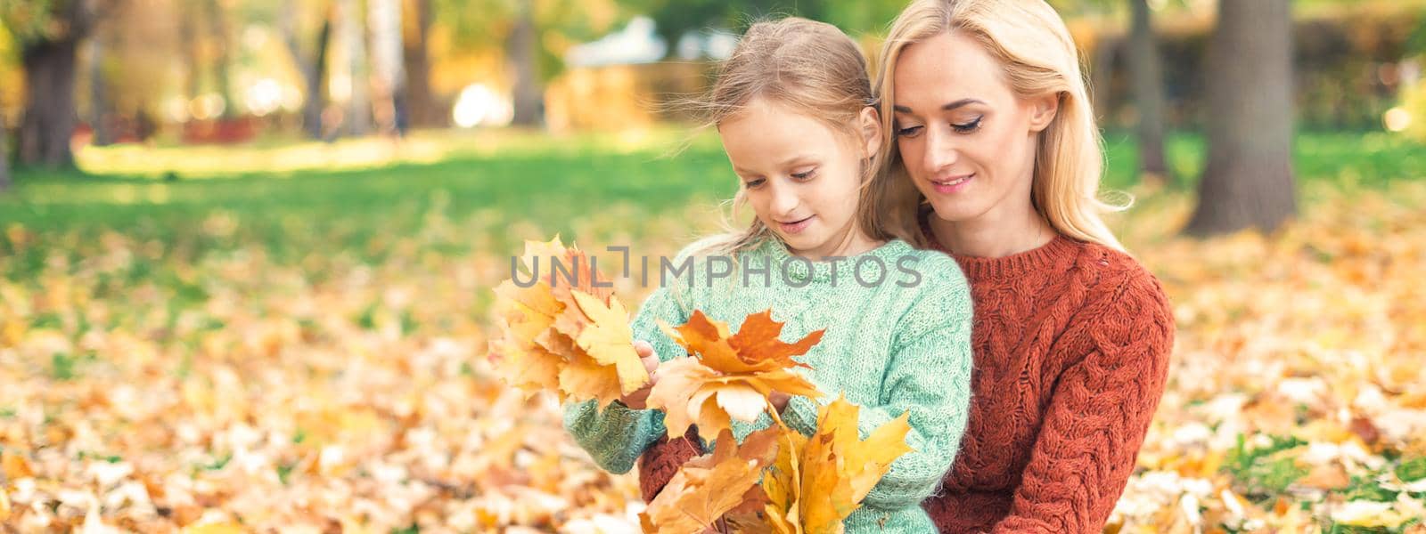 Happy young caucasian woman and little girl holding autumn yellow leaves sitting at the park