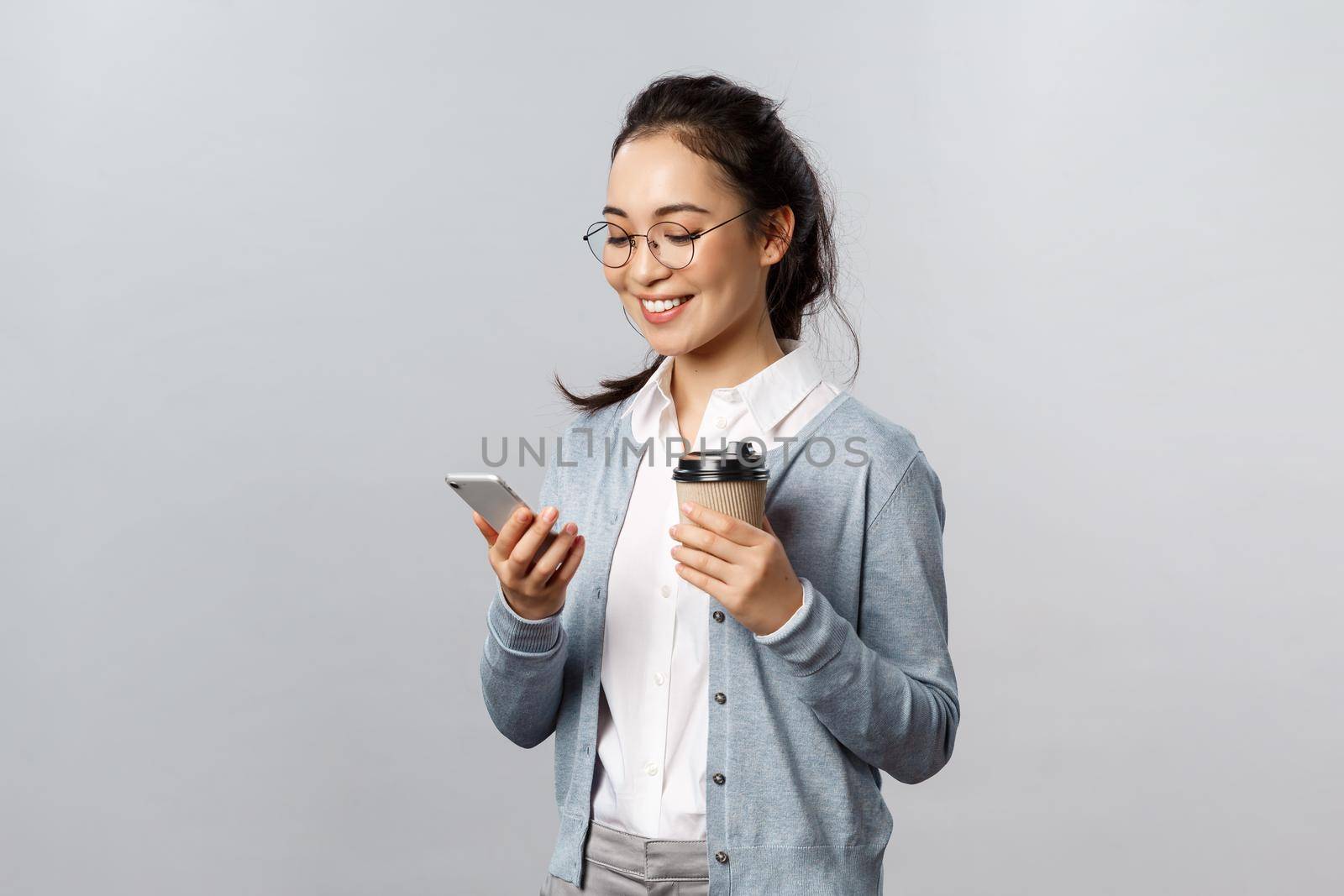 Office lifestyle, business and people concept. Attractive young korean woman checking her phone messages, drinking take-away coffee, look at smartphone display with pleased smile, texting in chat.