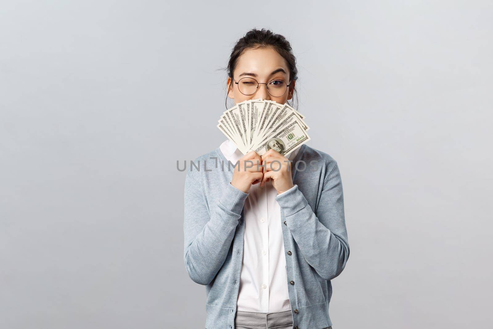 Business, finance and investment concept. Happy, pleased pretty asian woman, rich girl playfully raise eyebrow hiding satisfied smile behind money dollars, win cash prize, grey background.