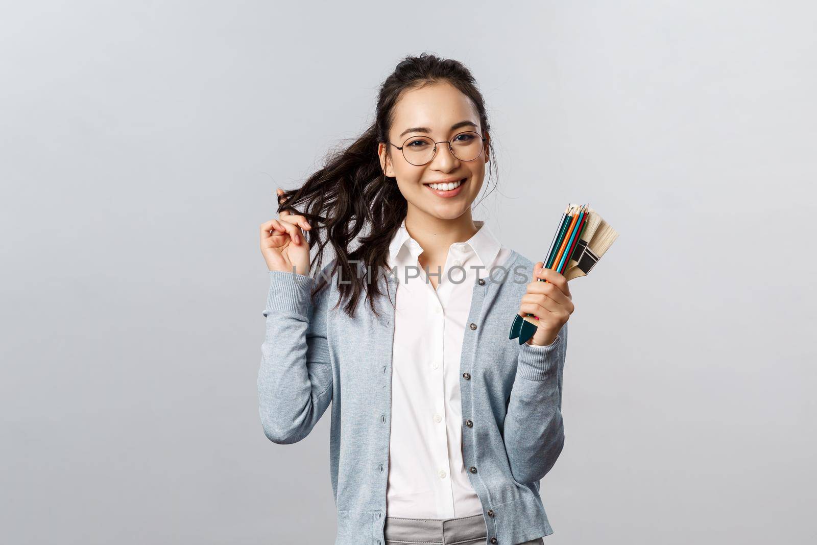 Hobbies, creativity and art concept. Cheerful, talented and creative cute asian girl in glasses, playing flirty with hair and smiling, holding colored pencils and painting brushes, grey background.