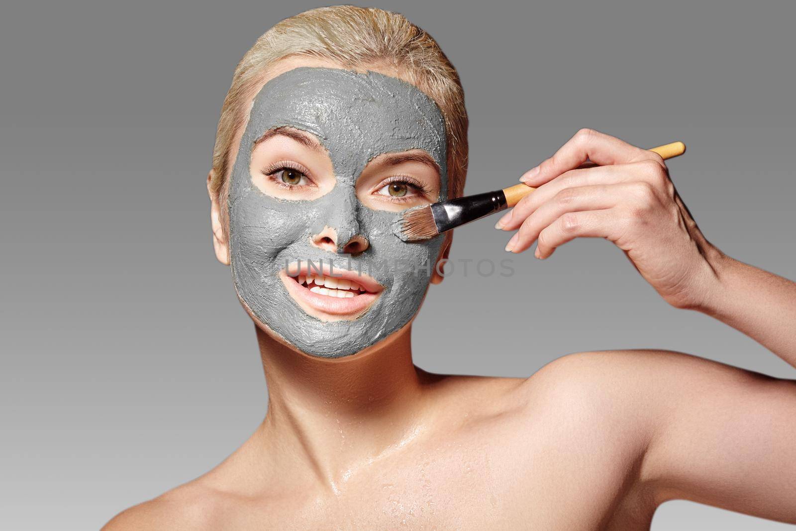 Beautiful Woman Applying White Facial Mask. Beauty Treatments. Close-up Portrait of Spa Girl Apply Clay Facial mask with Brush on grey background