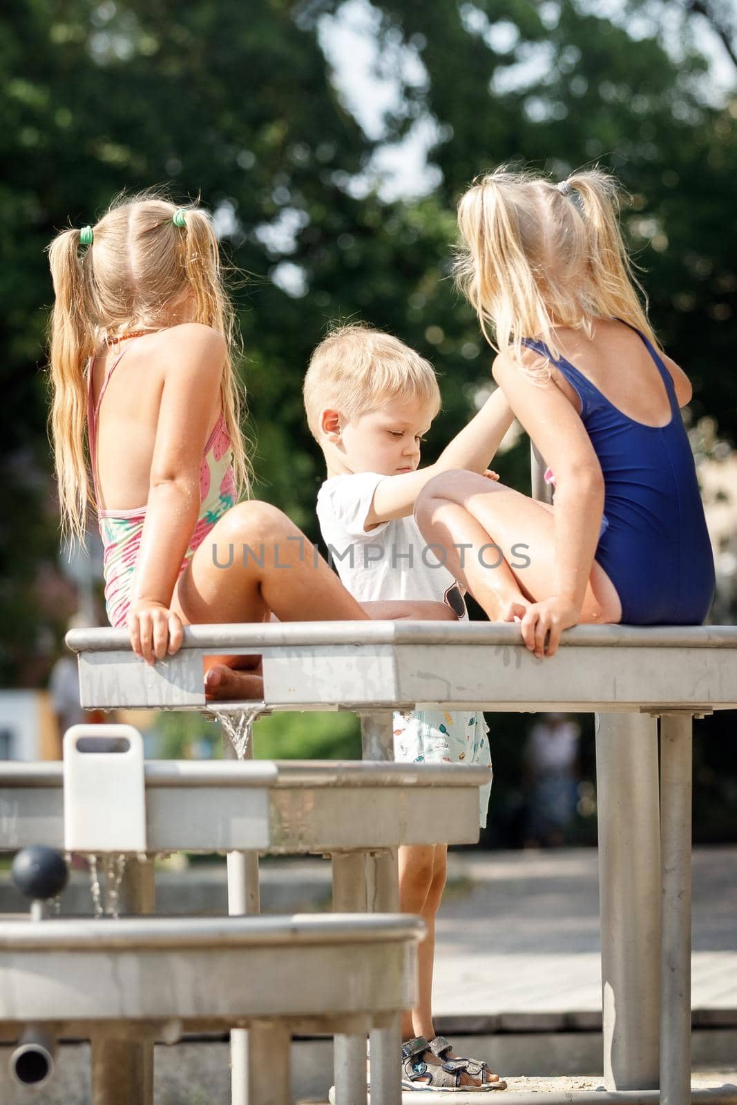 A happy boy plays and pumps water with special water equipment for children to play outdoors on a hot summer day. Two of his girlfriends are bathing in a fountain in a blue and pink bathing suit. by Lincikas