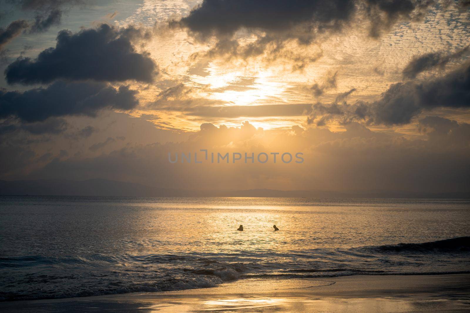 silhouette shot of people swimming under the golden sunlight god rays with clouds surrounding the sun and the blue gold water with waves lapping the shore showing peaceful life at havelock andaman islands by Shalinimathur