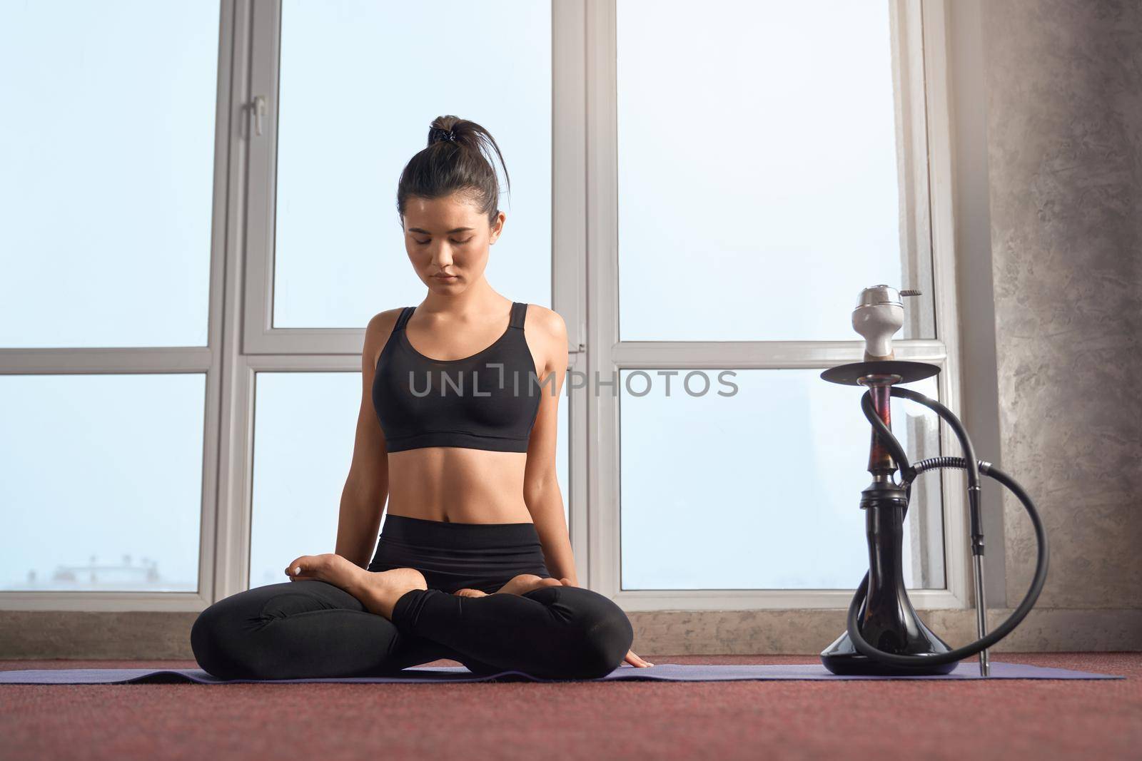 Front view of peaceful female yogi in black top and leggings meditating in lotus position on mat in studio. Woman with closed eyes practising yoga, with hookah on background. Concept of meditation.