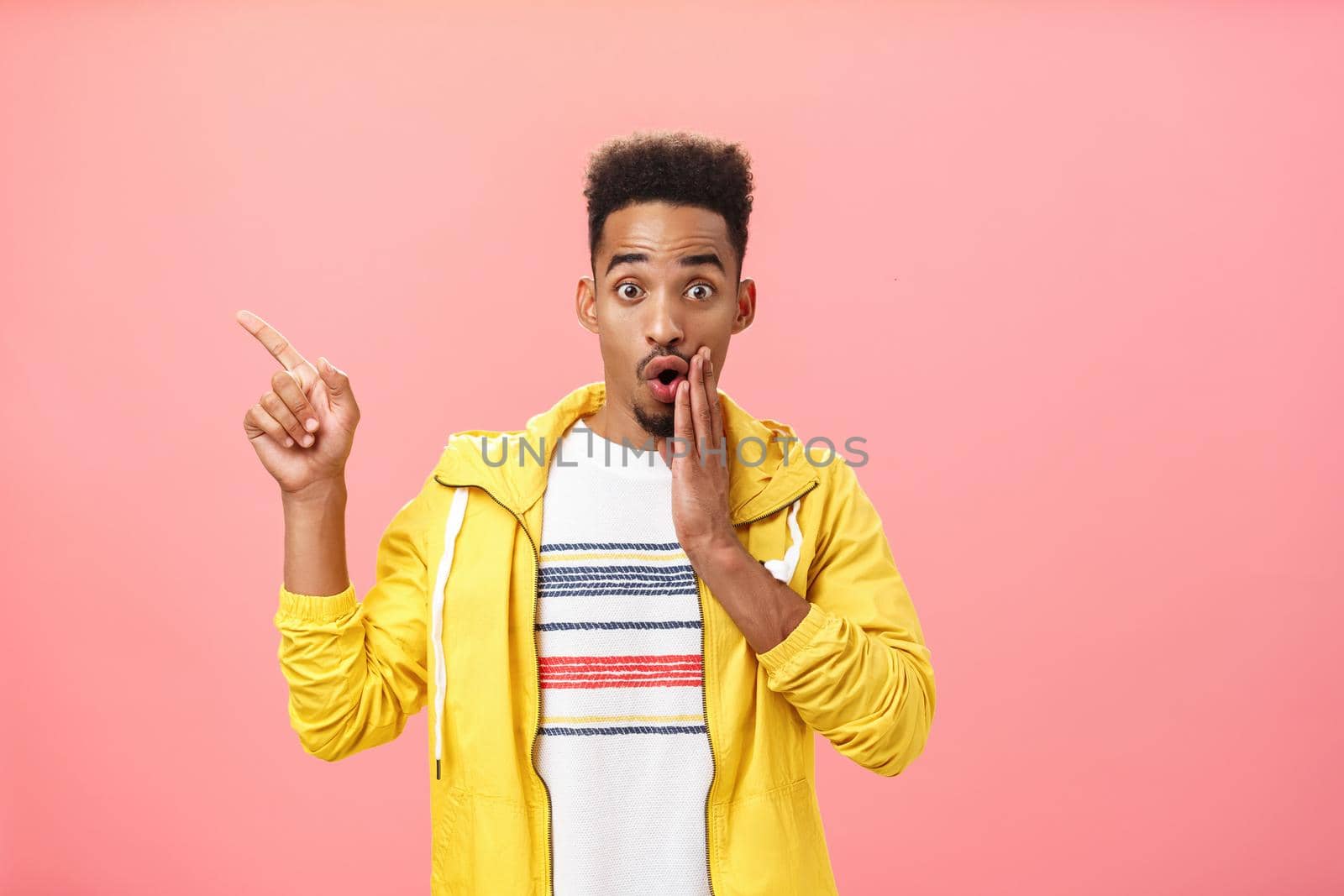 Look I am stunned. Portrait of impressed excited good-looking young male model in stylish yellow jacket opening mouth with gasp covering it with palm pointing amazed at upper left corner. Emotions and advertising concept