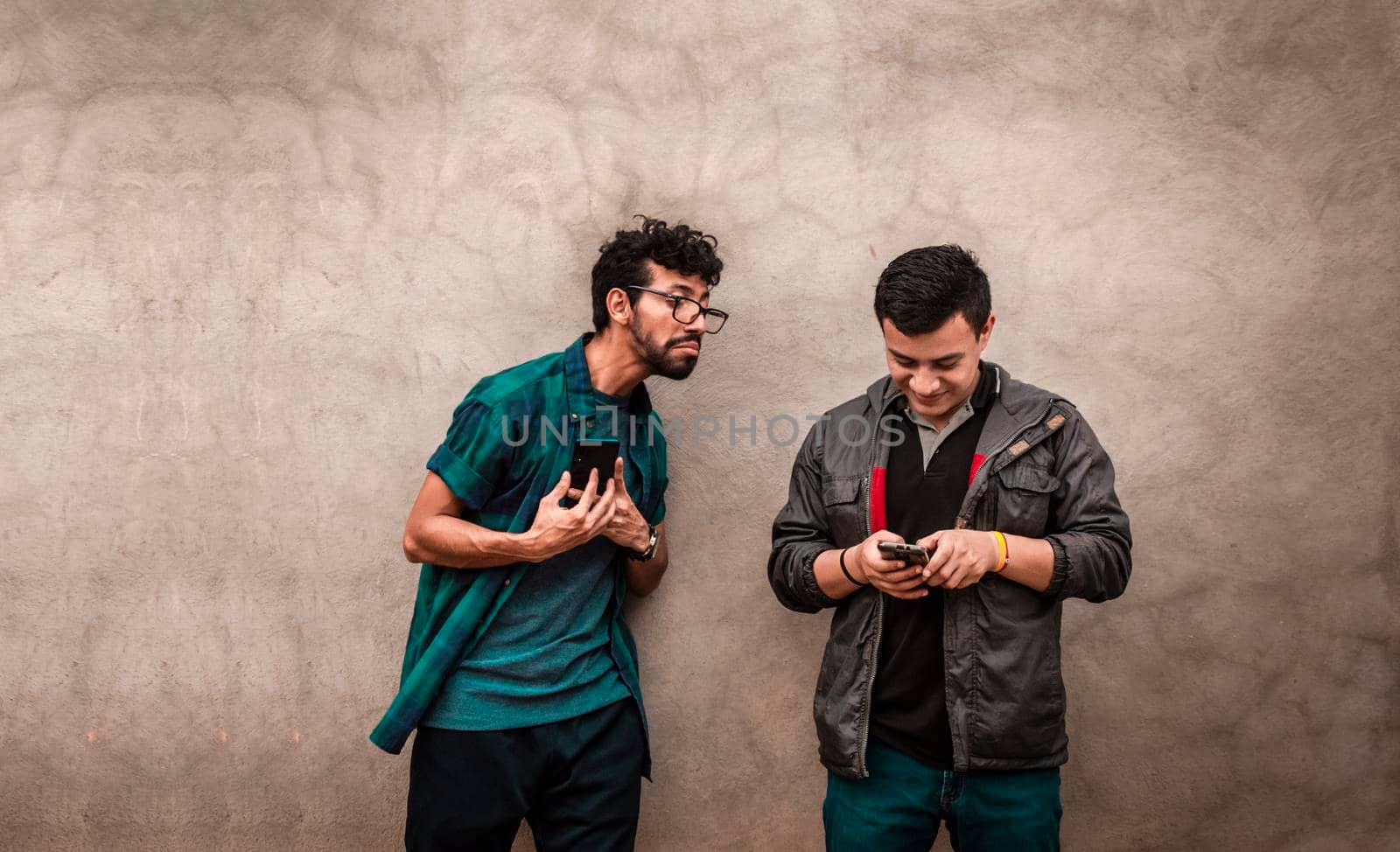 Young latin man spying on another cell phone, man showing his cell phone to another guy