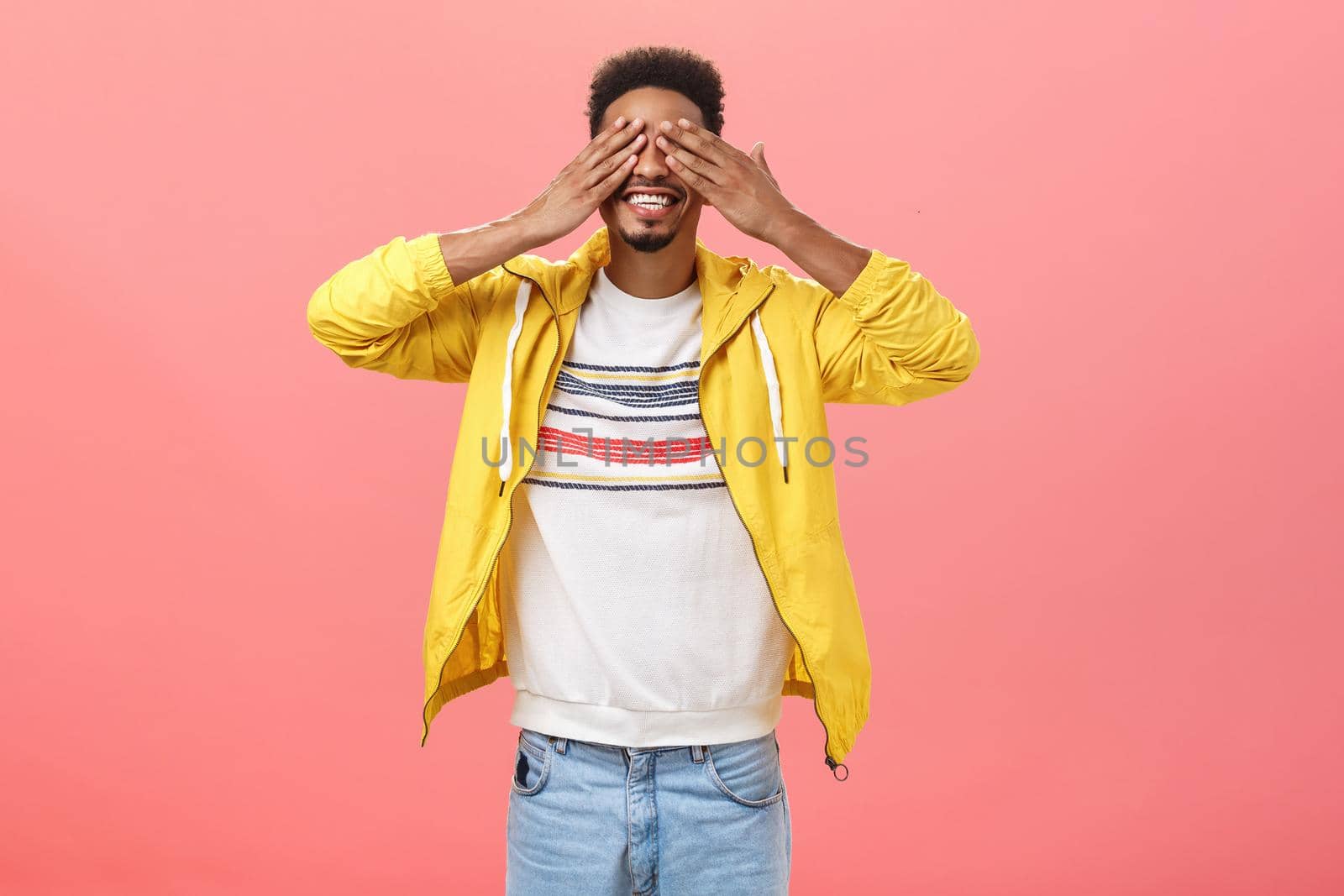 Can I open eyes. Awaiting happy and amused charming dark-skinned boyfriend with beard in trendy yellow jacket closing sight with palms on face smiling broadly waiting for surprise over pink wall. Body language concept