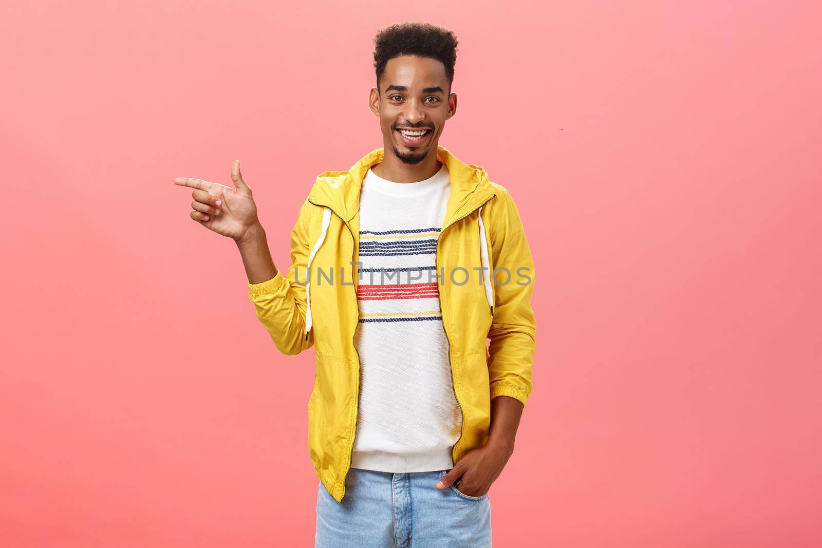 Friendly helpful male dark-skinned friend showing girlfriend appartment pointing left at living room smiling joyfully hanging out in funny and amusing company over pink background. Emotions and directions concept