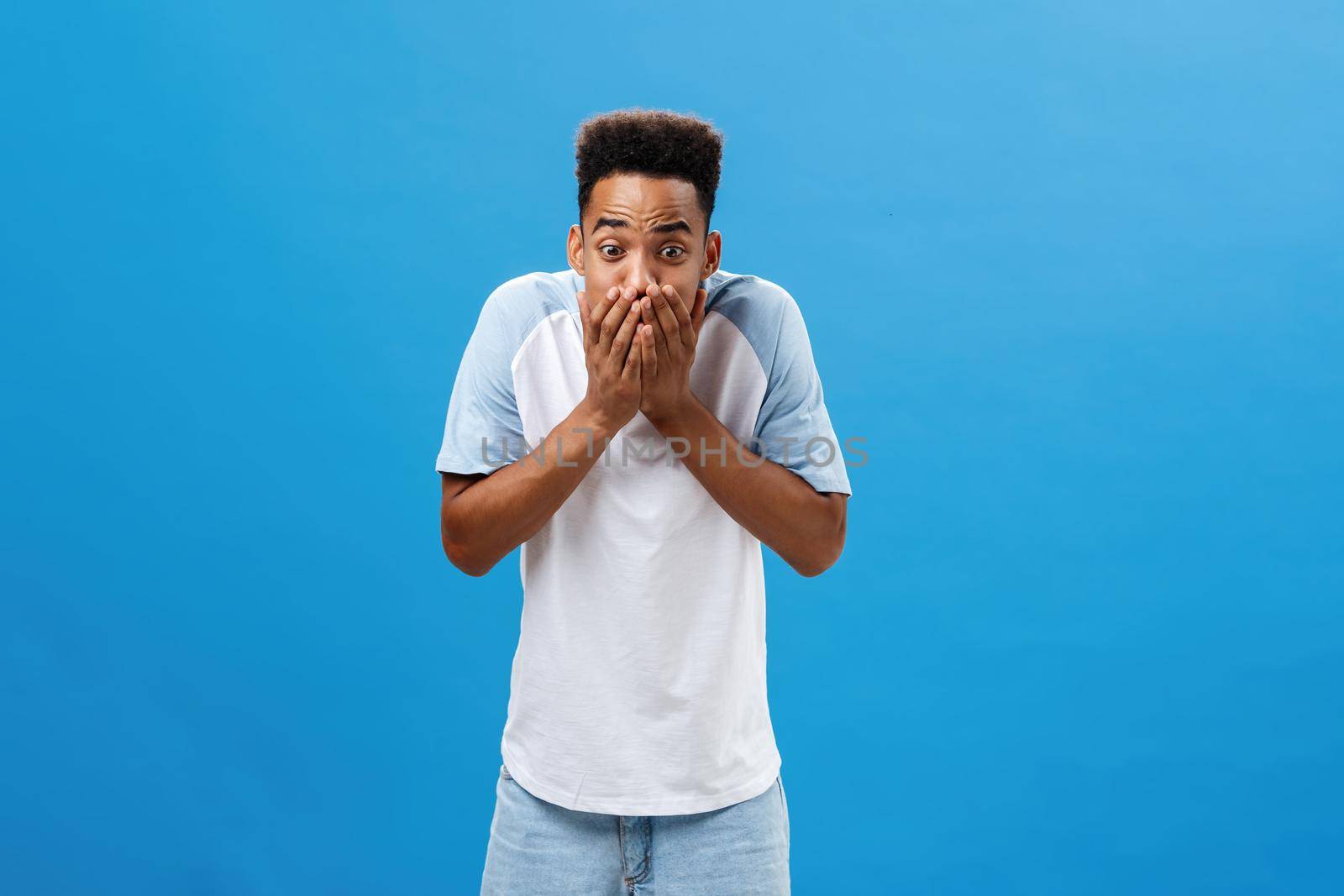 Shocked young dark-skinned male student witnessing murder screaming and starting panic covering mouth with both palms to calm down raising eyebrows staring with fear and fright over blue wall. Emotions concept