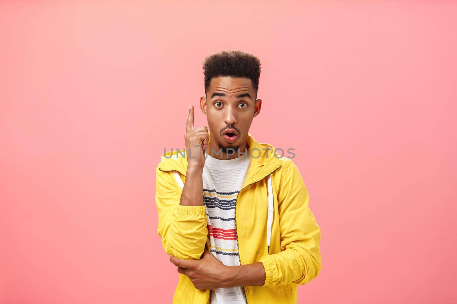 Got it finally understood. Portrait of excited smart dark-skinned creative male with beard in trendy yellow jacket raising index finger in eureka gesture folding lips adding suggestion over pink wall.