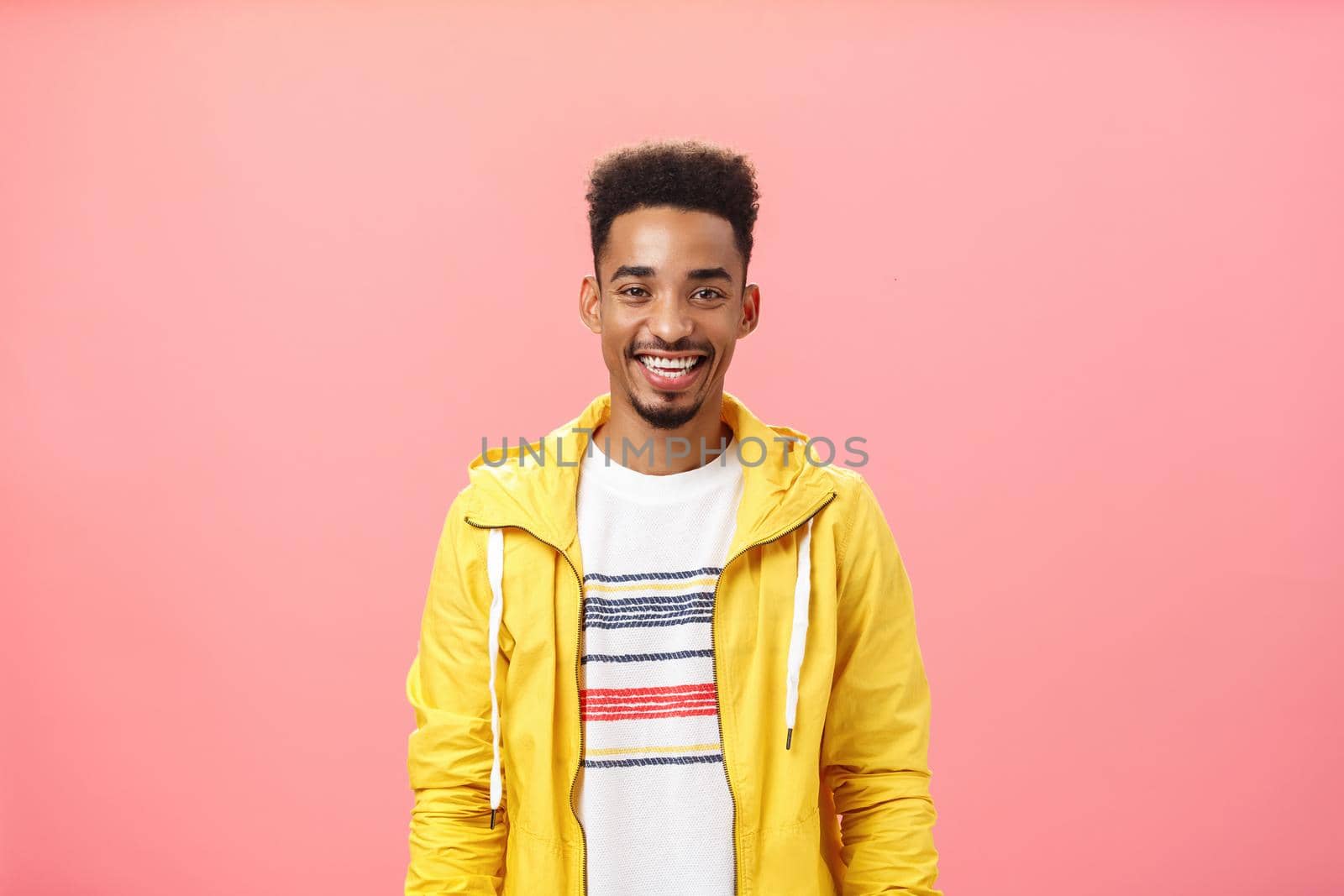Waist-up shot of charming friendly good-looking dark-skinned male with beard and afro hairstyle in trendy yellow jacket smiling with satisfied pleased grin standing over pink background. Emotions concept