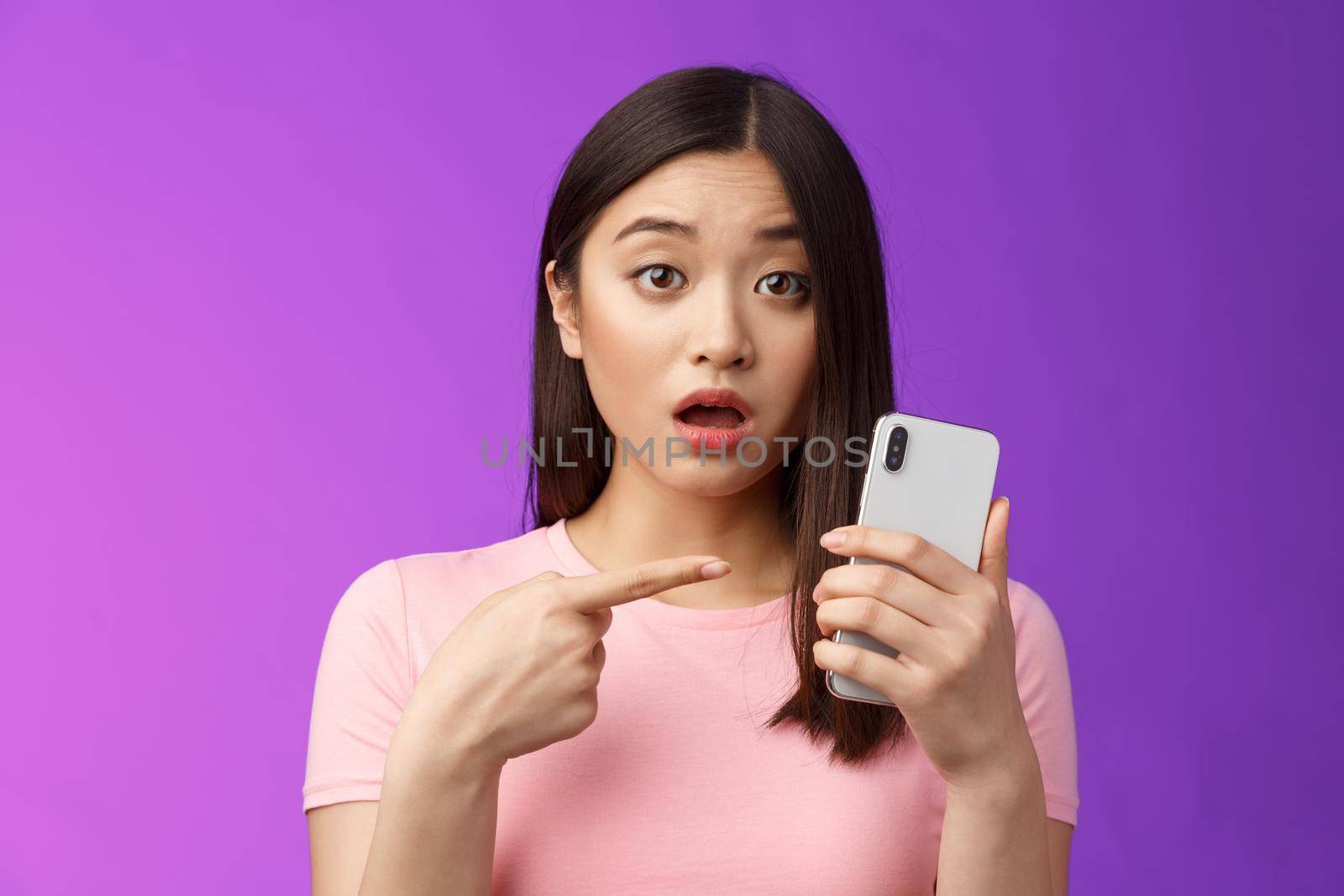 Concerned confused cute young asian woman hold smartphone open mouth, pointing finger telephone, look questioned upset, troubled see strange post online, stand purple background by Benzoix
