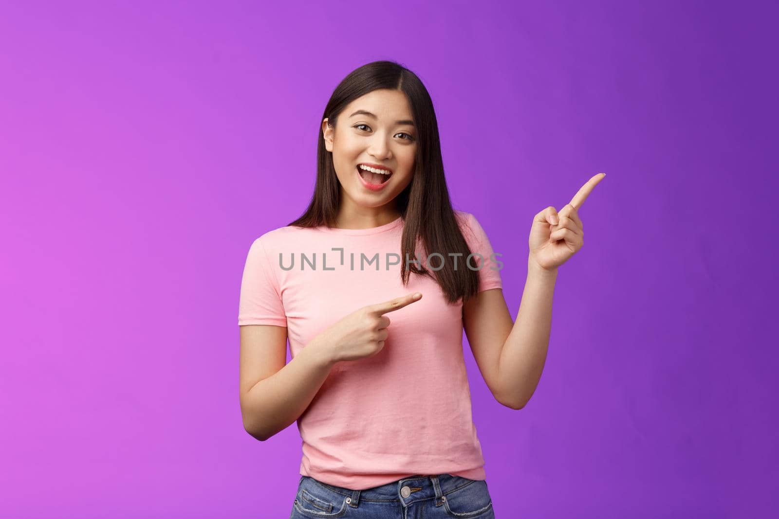 Friendly enthsuasitic amazed attractive tender asian girl having interesting conversation, discuss interest new store, pointing right inviting come inside, stand purple background smiling camera.
