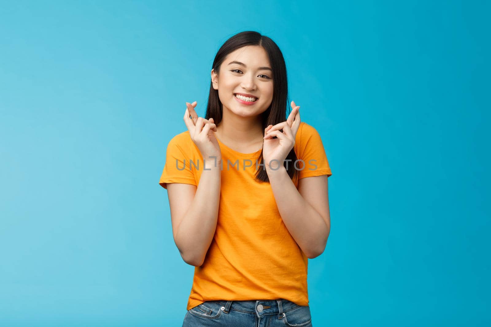 Cheerful smiling hopeful asian girl praying wish for good luck, cross fingers grinning toothy anticipate positive good news, stand blue background supplicating optimistic feelings hope win.