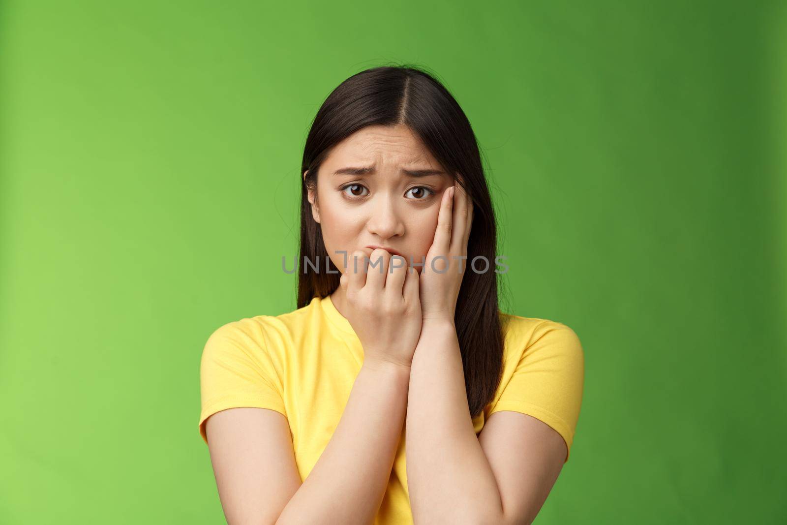 Innocent insecure timid asian scared girl panicking, standing afraid victim terrified, touch cheek shocked, frowning stunned, biting fingernails, anxiously stare camera, green background by Benzoix