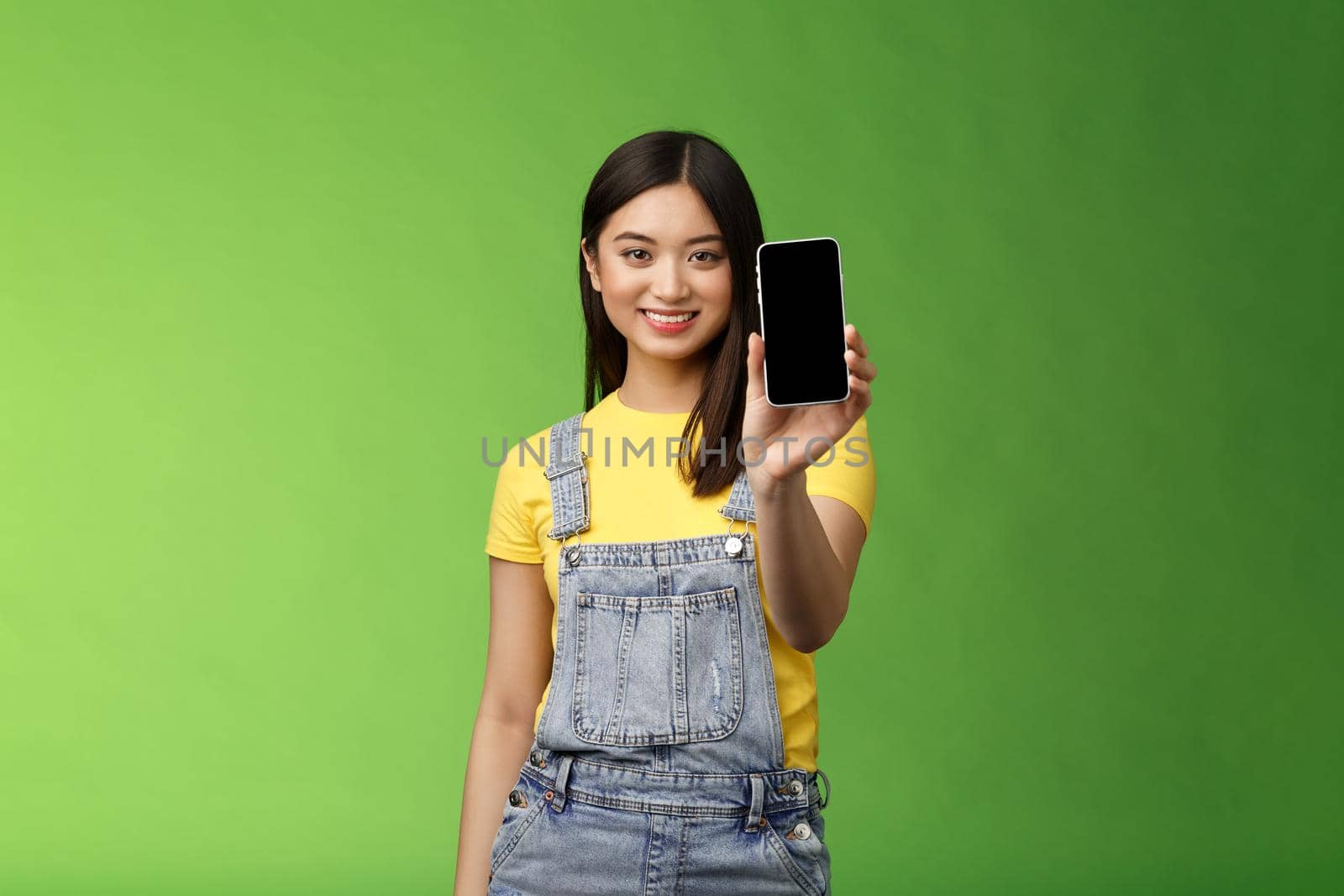 Confident assertive good-looking female asian introduce smartphone app, extend hand hold telephone arm, smiling delighted, recommend social profile, bragging own blog, green background.