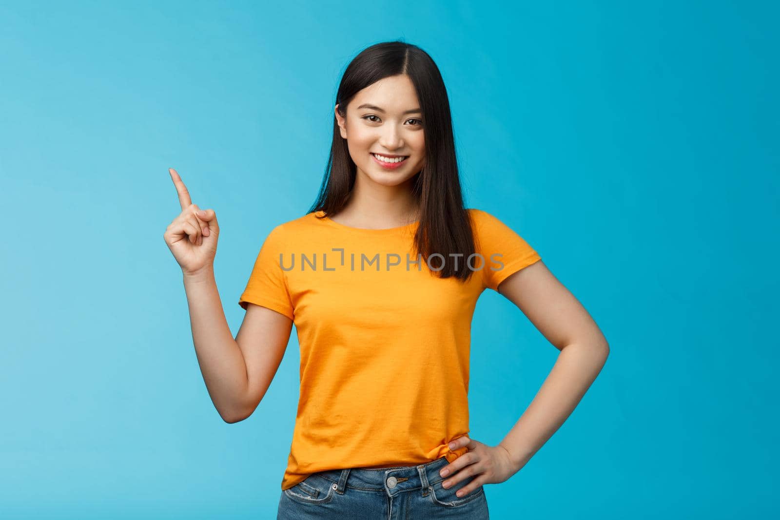 Happy confident asian girl self-assured pointing upper left corner, show way, smiling determined, assertive introduce online store promo, stand blue background in yellow t-shirt. Copy space