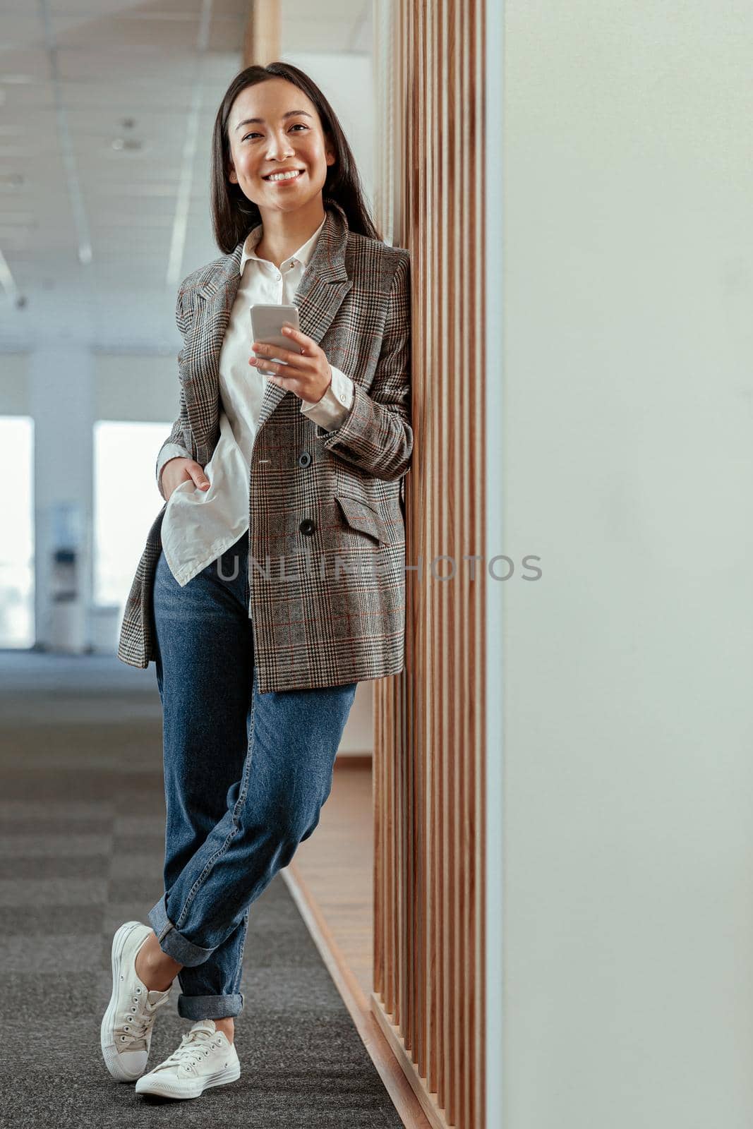 Portrait of smiling asian business woman standing in modern office with phone by Yaroslav_astakhov