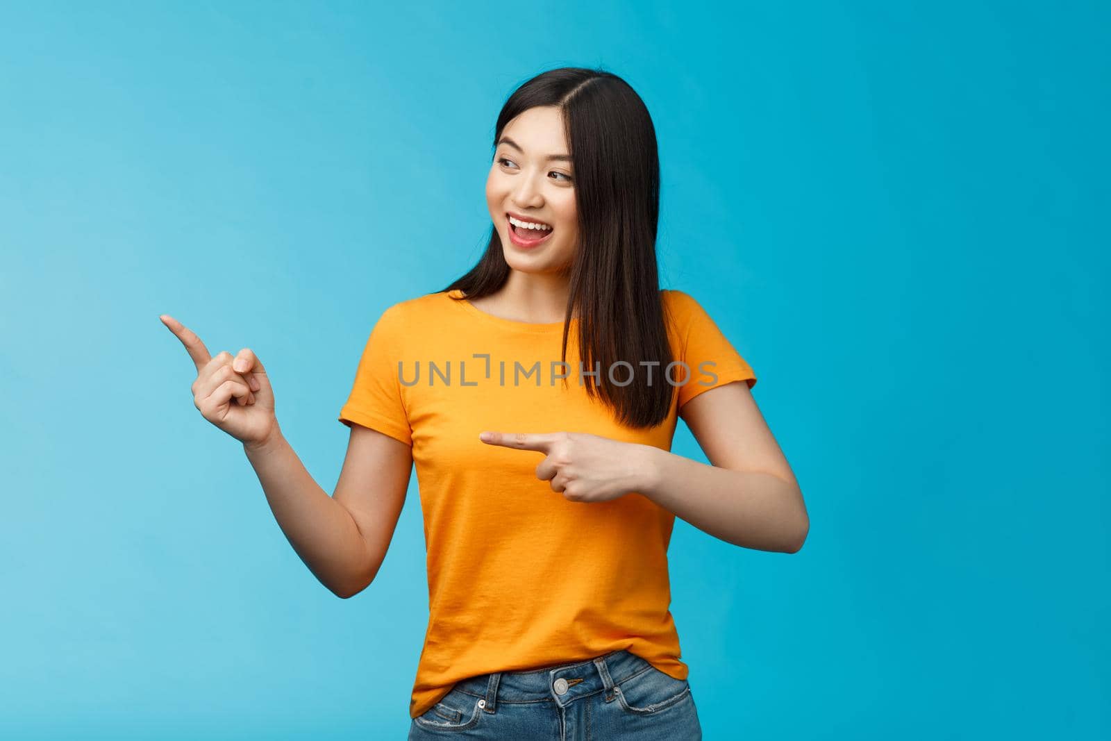 Friendly enthusiastic asian girl curiously looking sideways, pointing left interesting object, grinning joyfully discuss new opened store, stand blue background carefree lively talking.
