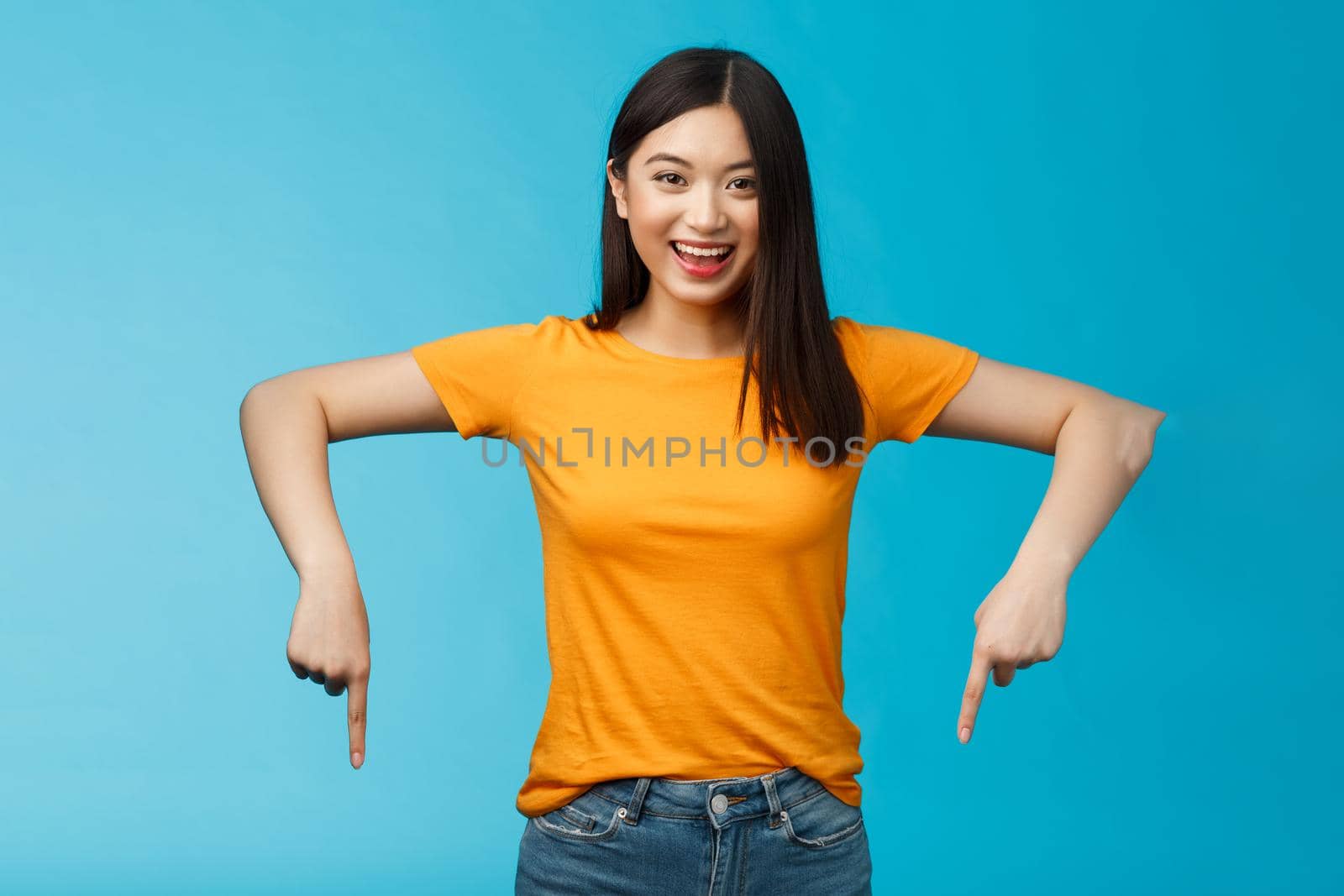 Enthusiastic pleasant asian blong modern female student inviting you visit awesome event pointing down indicating bottom advertisement, stand blue background smiling broadly look happily camera.