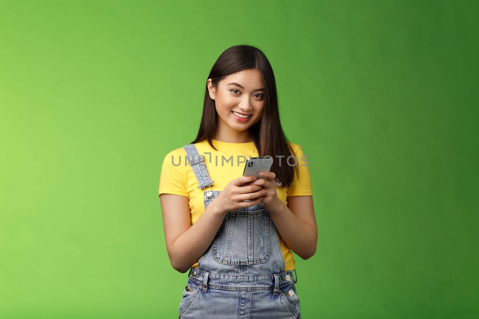 Cute tender asian gorgeous woman using smartphone, smiling carefree, look camera, texting friend, waiting boyfriend finish work, stand cafe green background, hold telephone, order food online by Benzoix